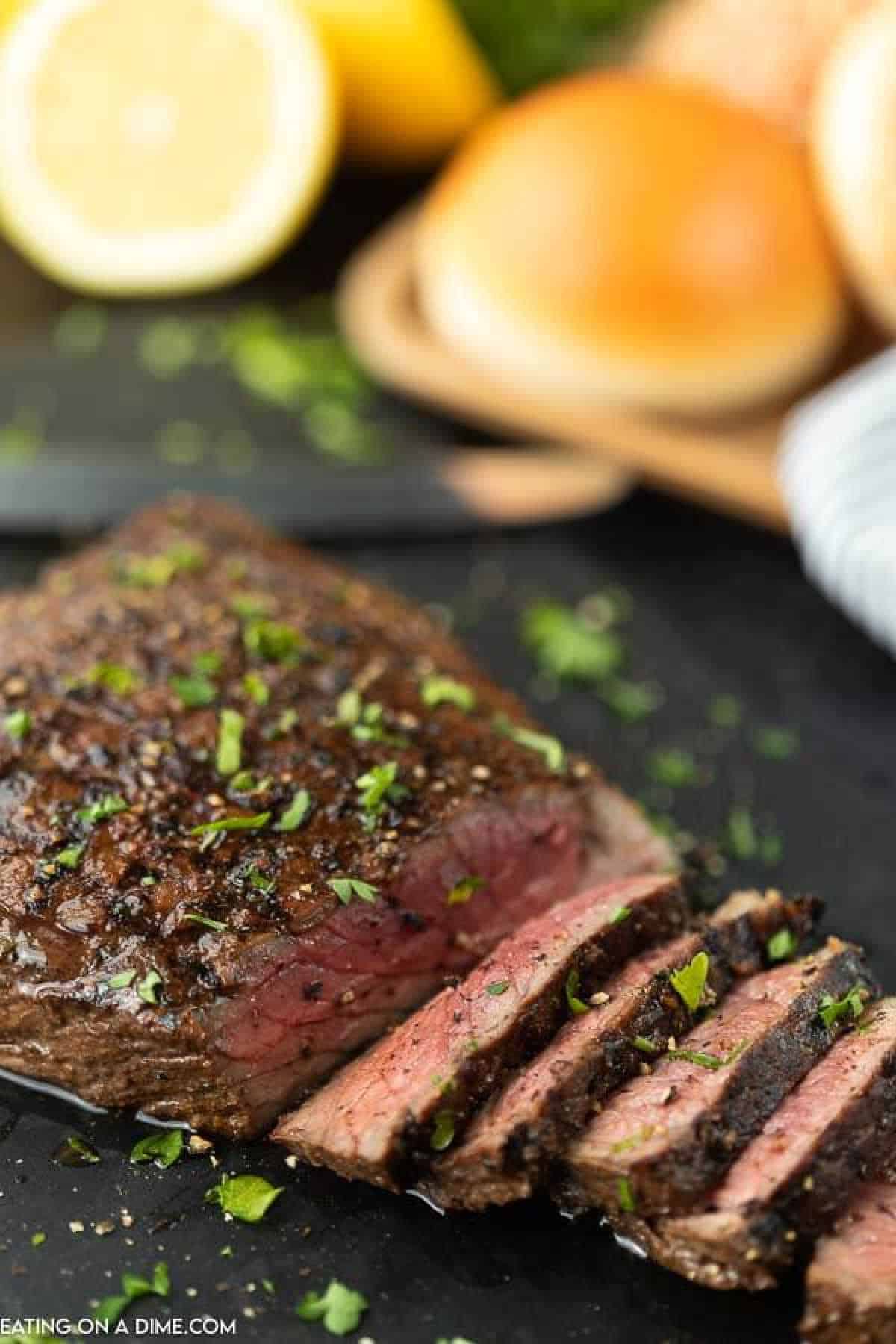 Close-up of a sliced, juicy steak seasoned with a rich London broil marinade and garnished with chopped herbs. There are lemons and bread rolls in the background, placed on a sleek black surface.