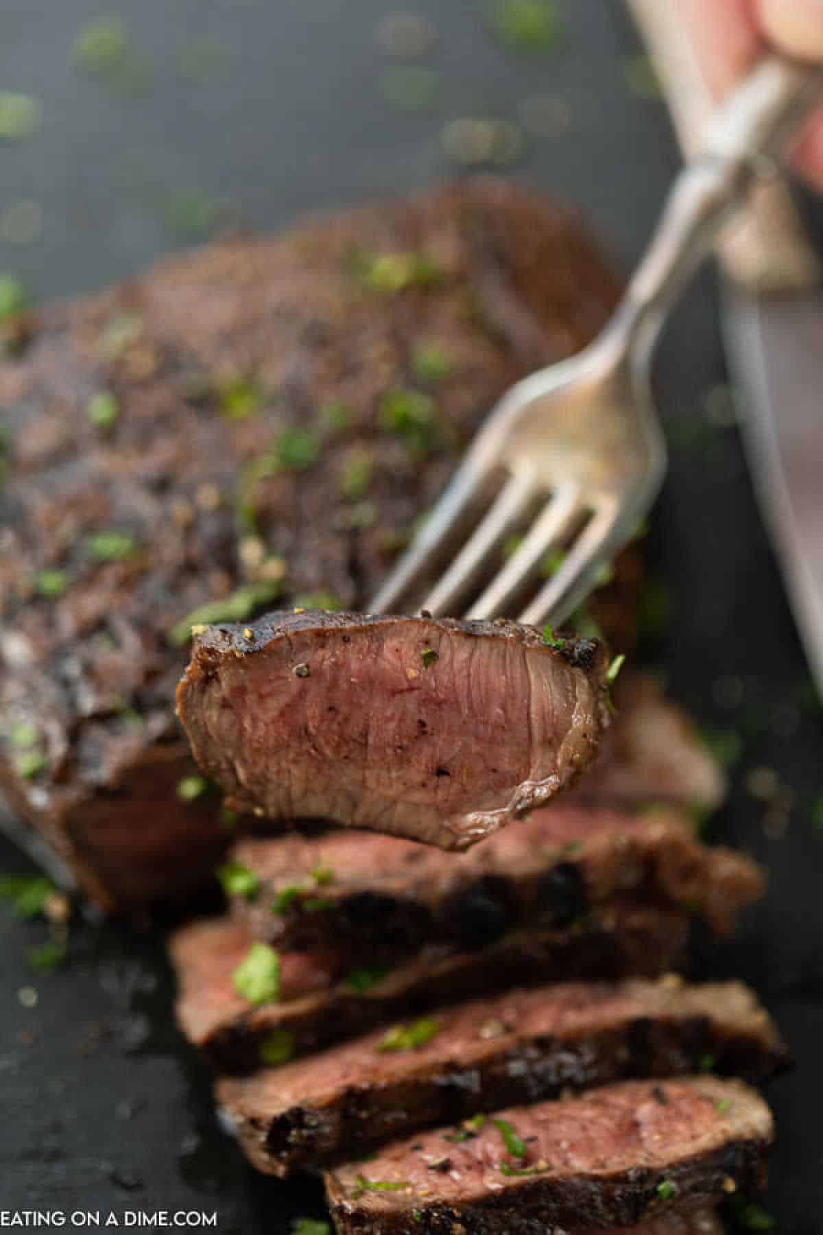 A fork holding a slice of medium-cooked steak marinated in a rich London broil marinade showcases its tender, juicy center. The steak is garnished with chopped herbs and lies on a cutting board, with several neatly arranged slices in the background.