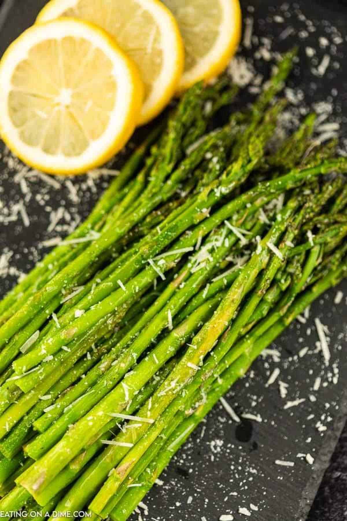 Oven-roasted green asparagus spears, topped with grated parmesan cheese, are exquisitely arranged on a black platter, with lemon slices in the background enhancing the dish's appeal.