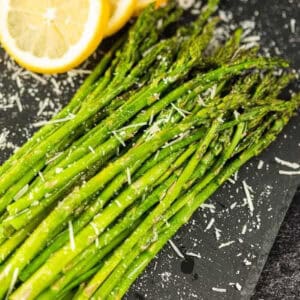 Oven-roasted parmesan asparagus spears are elegantly arranged on a black slate surface, their flavors elevated by the addition of grated Parmesan cheese. A few lemon slices rest in the top left corner, adding a vibrant touch to this delightful presentation.