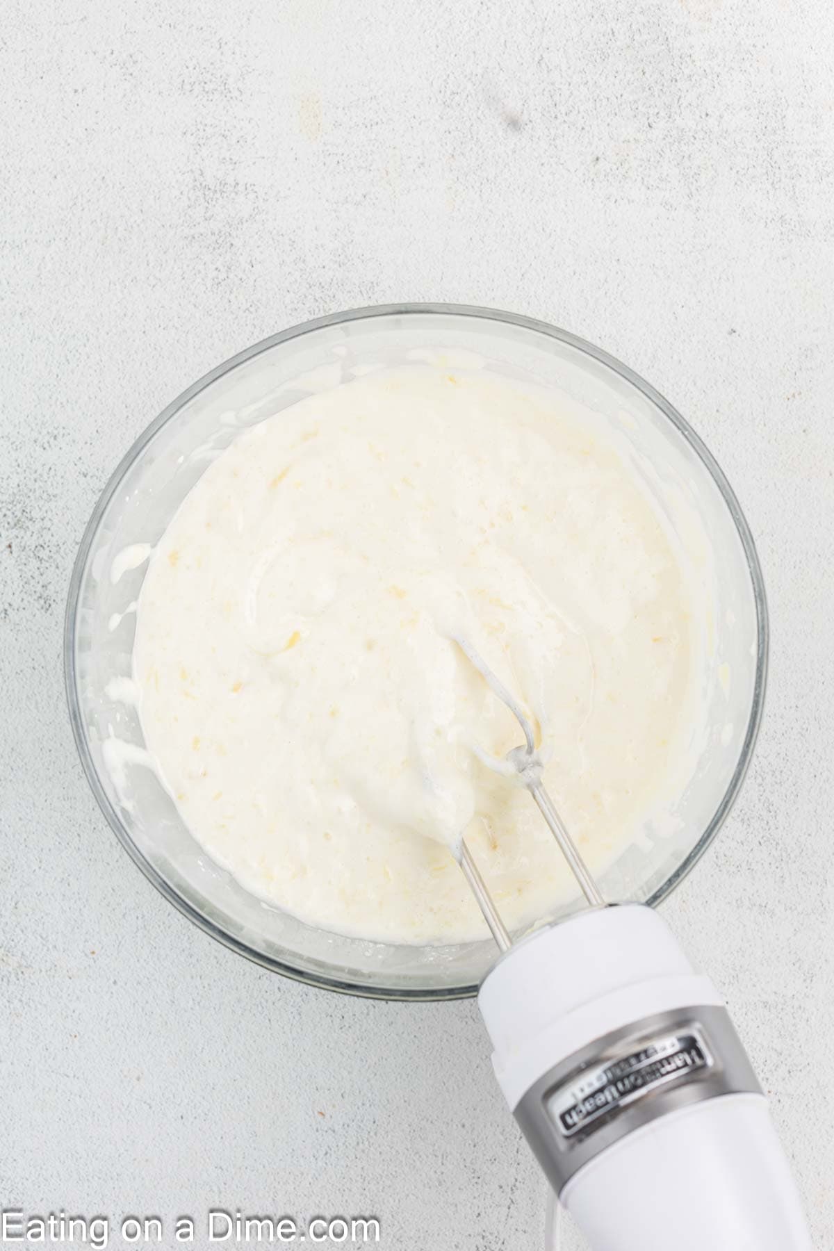 A white hand mixer whirs through creamy white batter for a Pineapple Angel Food Cake in a glass bowl on a light gray surface.