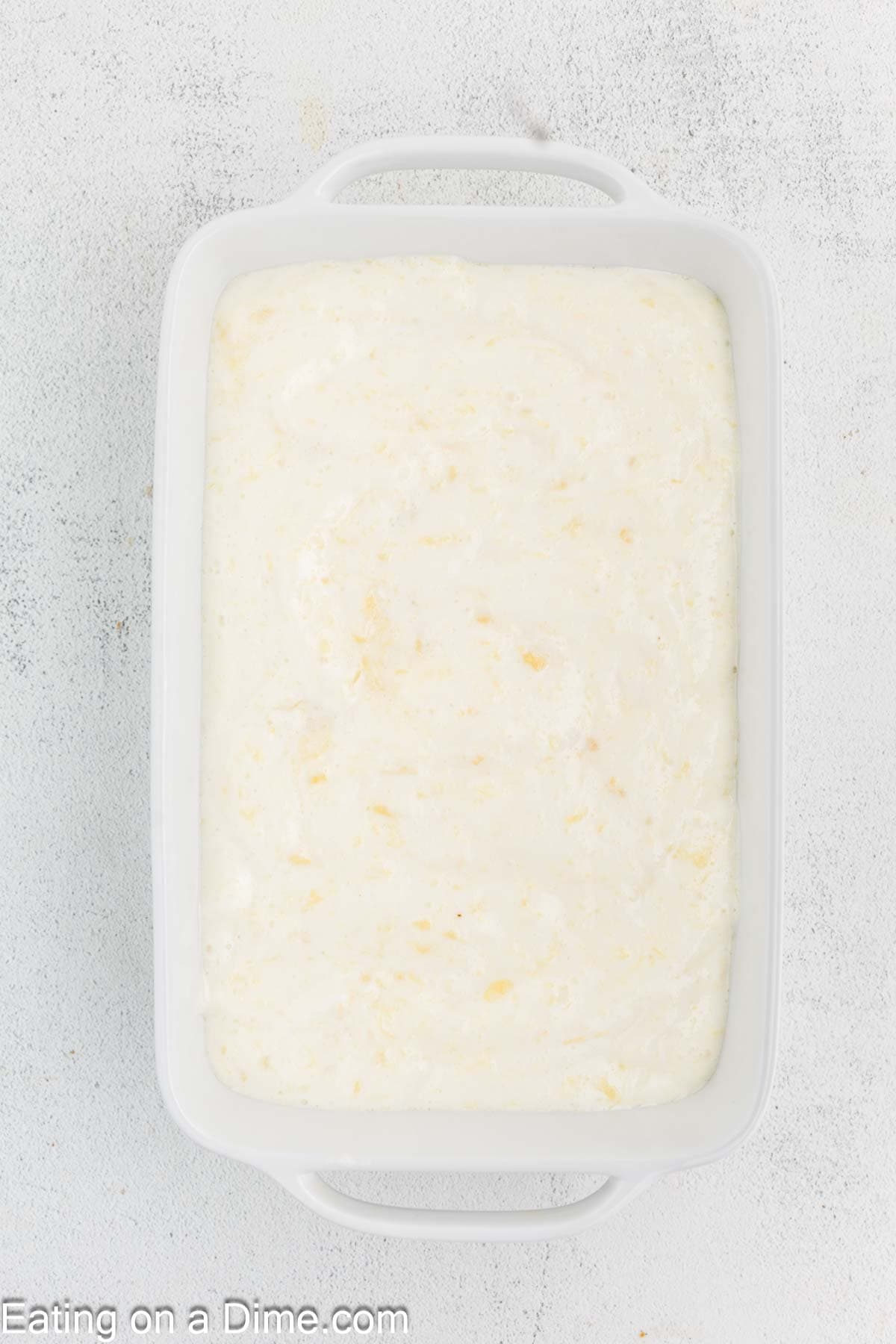 A rectangular white baking dish filled with a creamy Pineapple Angel Food Cake mixture rests on a light gray textured surface, exuding an inviting aroma perfect for dessert lovers.