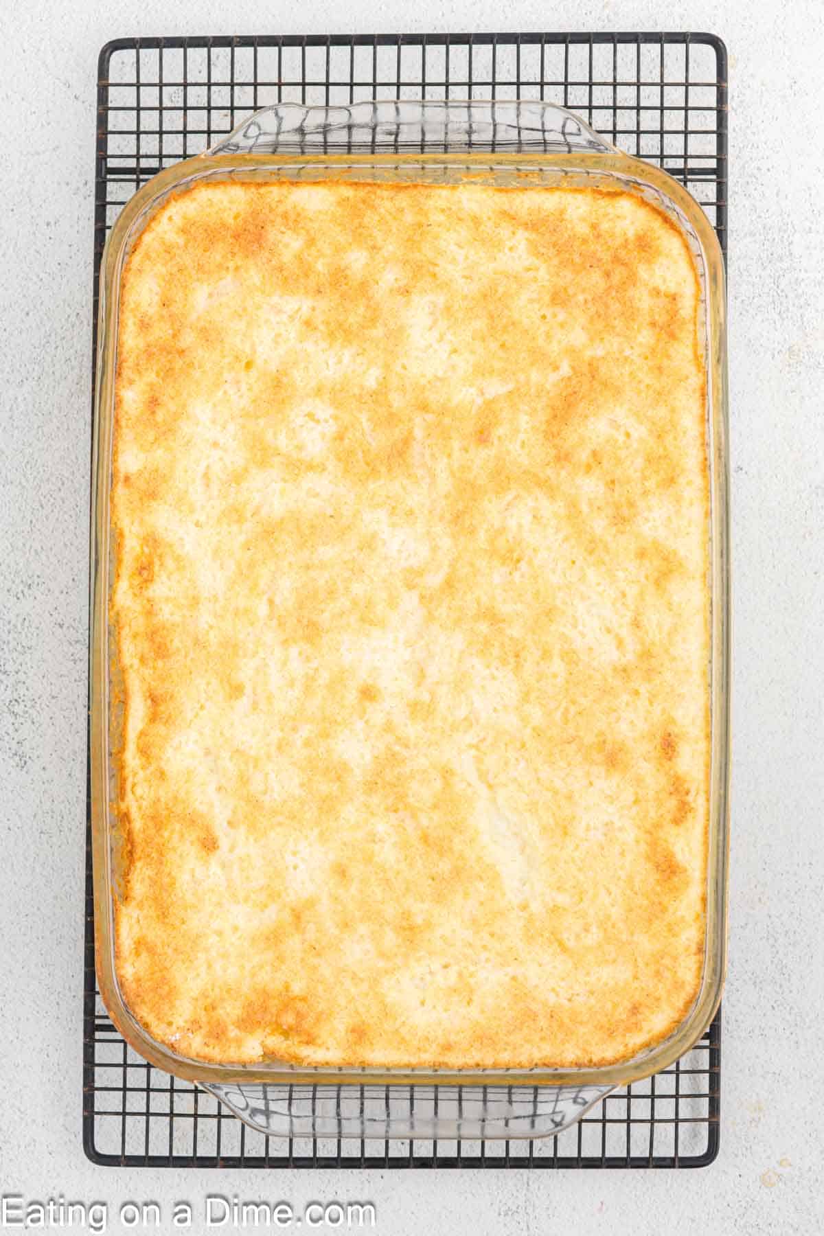 A golden-brown Pineapple Angel Food Cake rests in a rectangular glass pan on a black cooling rack. The surface is evenly browned, hinting at the delightful flavors inside. The light gray background complements the scene while "Eating on a Dime.com" is subtly noted at the bottom.