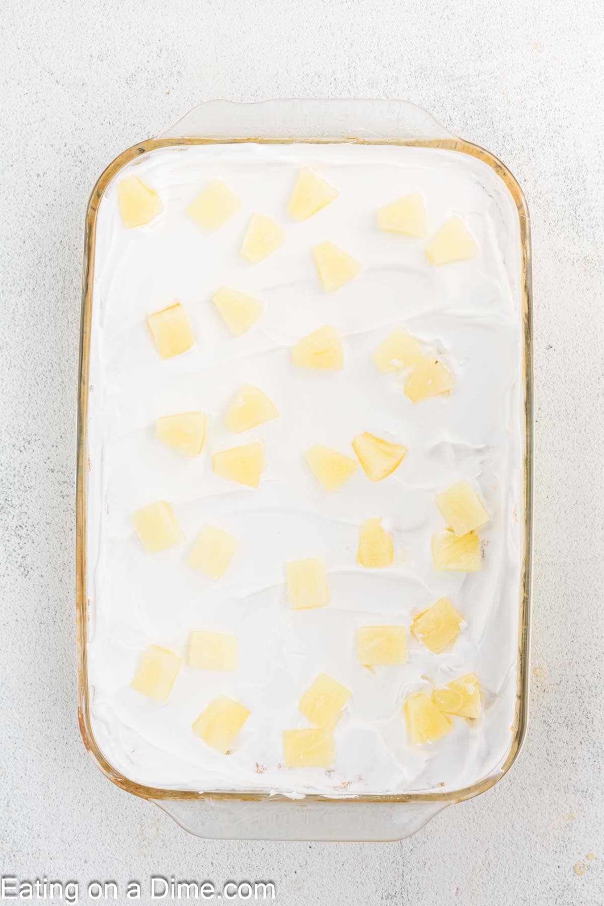 A rectangular glass dish cradles a delightful Pineapple Angel Food Cake, its surface swathed in white cream and adorned with evenly spaced chunks of pineapple. The ensemble rests against a speckled white backdrop, inviting both the eye and palate.