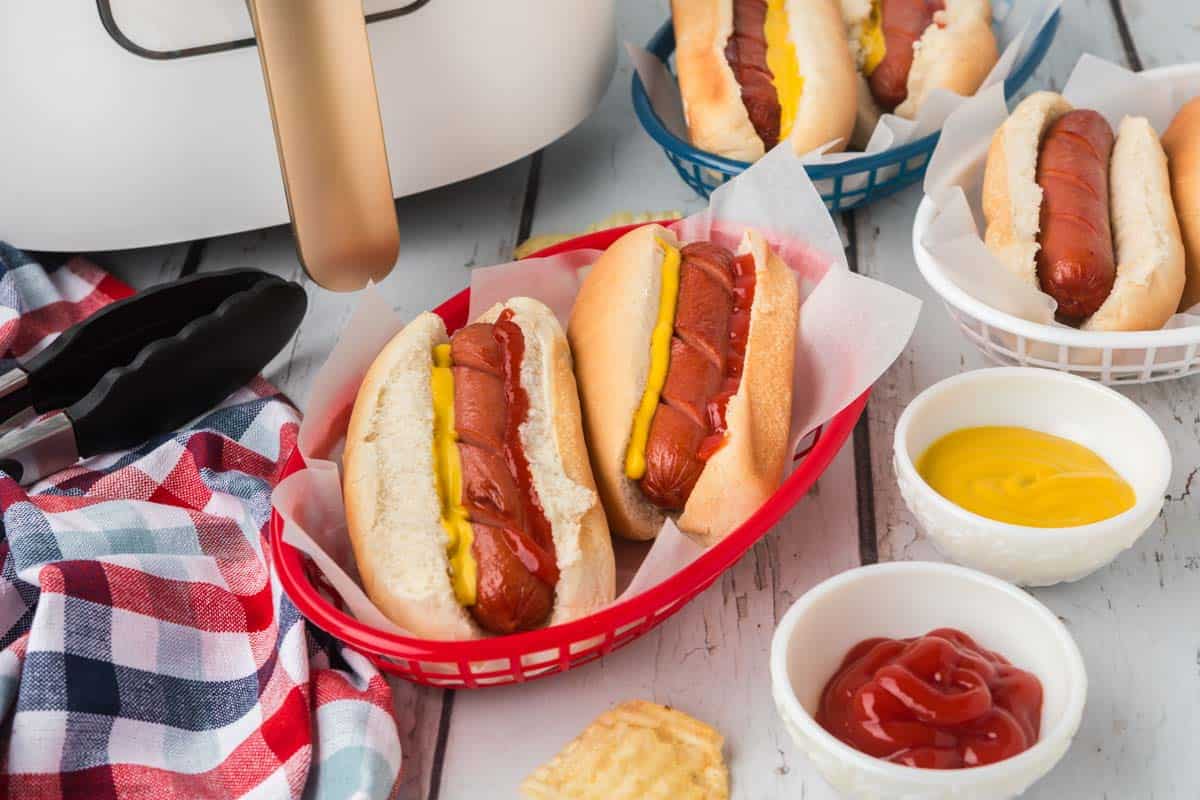 Hot dogs in a red basket topped with mustard and ketchup with a bowl of ketchup and mustard on the side