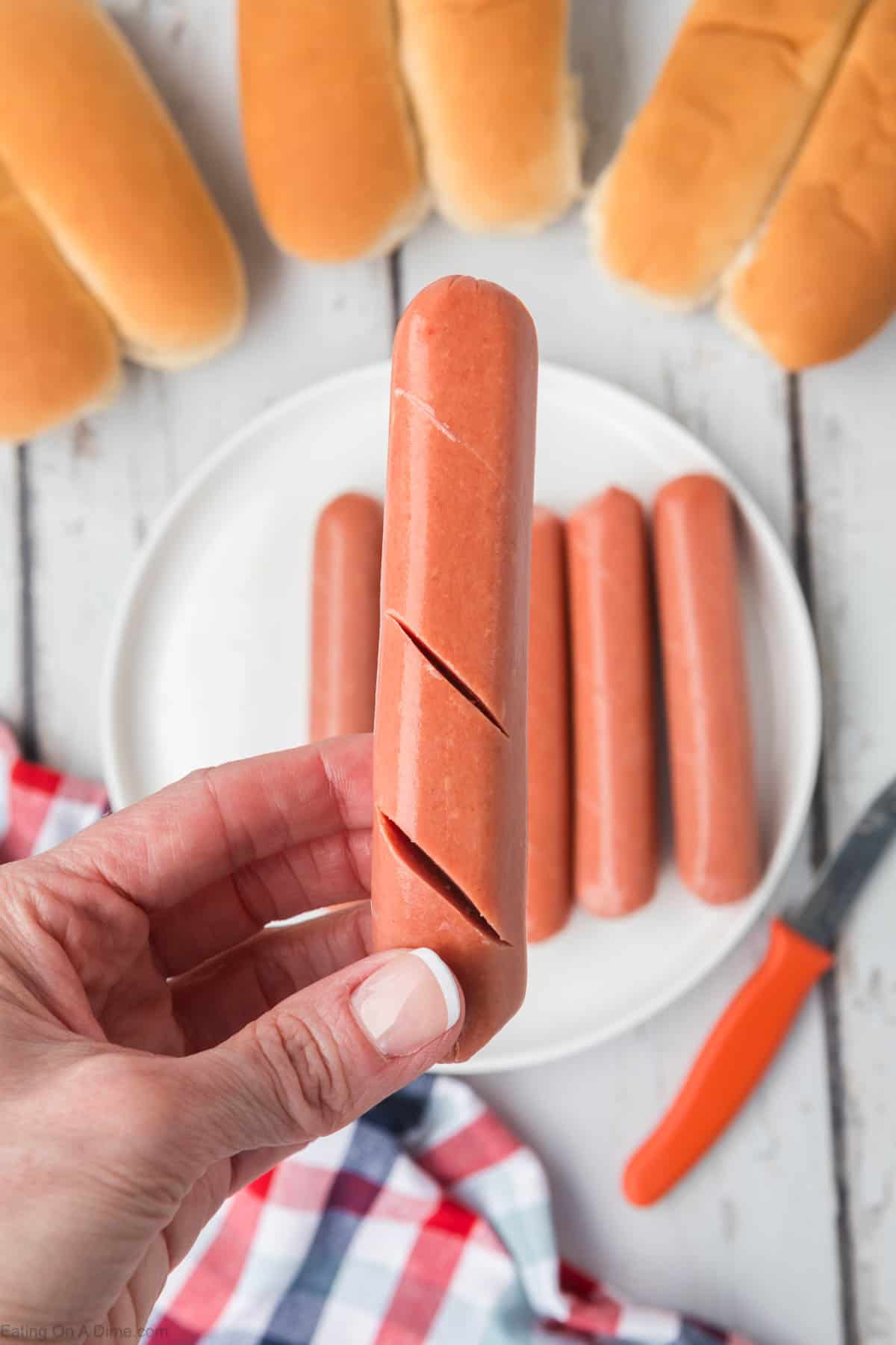 Hot dogs cut with slits with a plate of hot dogs and buns in the back ground