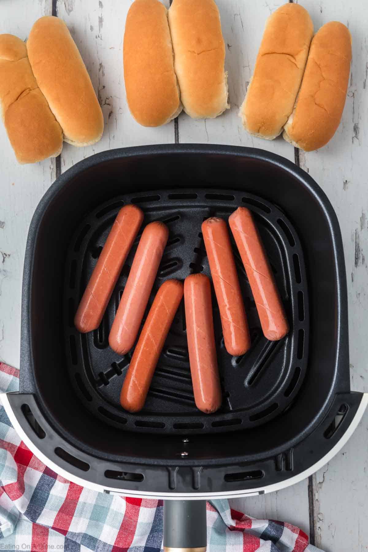 Hot dogs in the air fryer basket with a hot dogs buns on the side