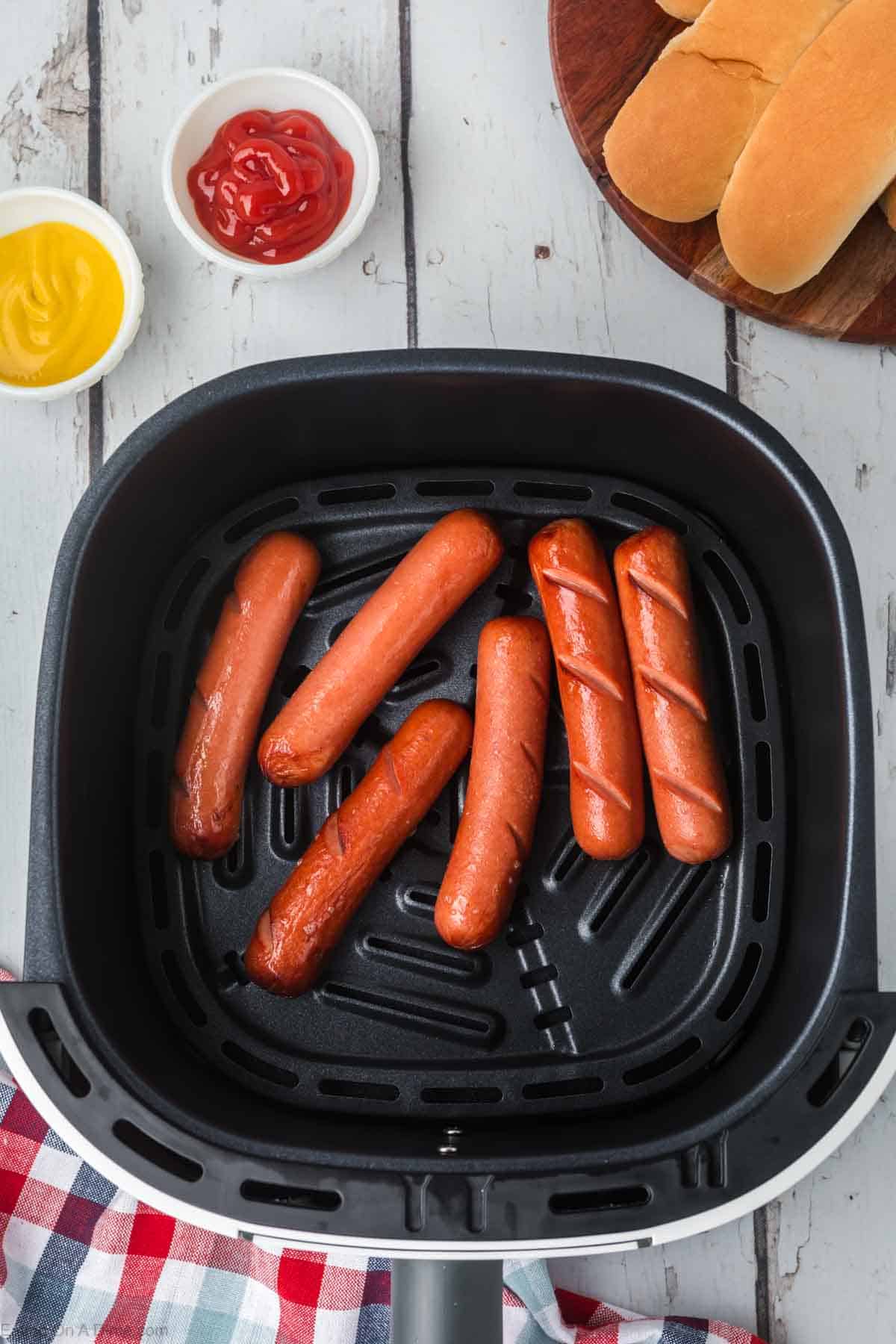 Cooked hot dogs in an air fryer basket with a bowl of mustard and ketchup on the side 