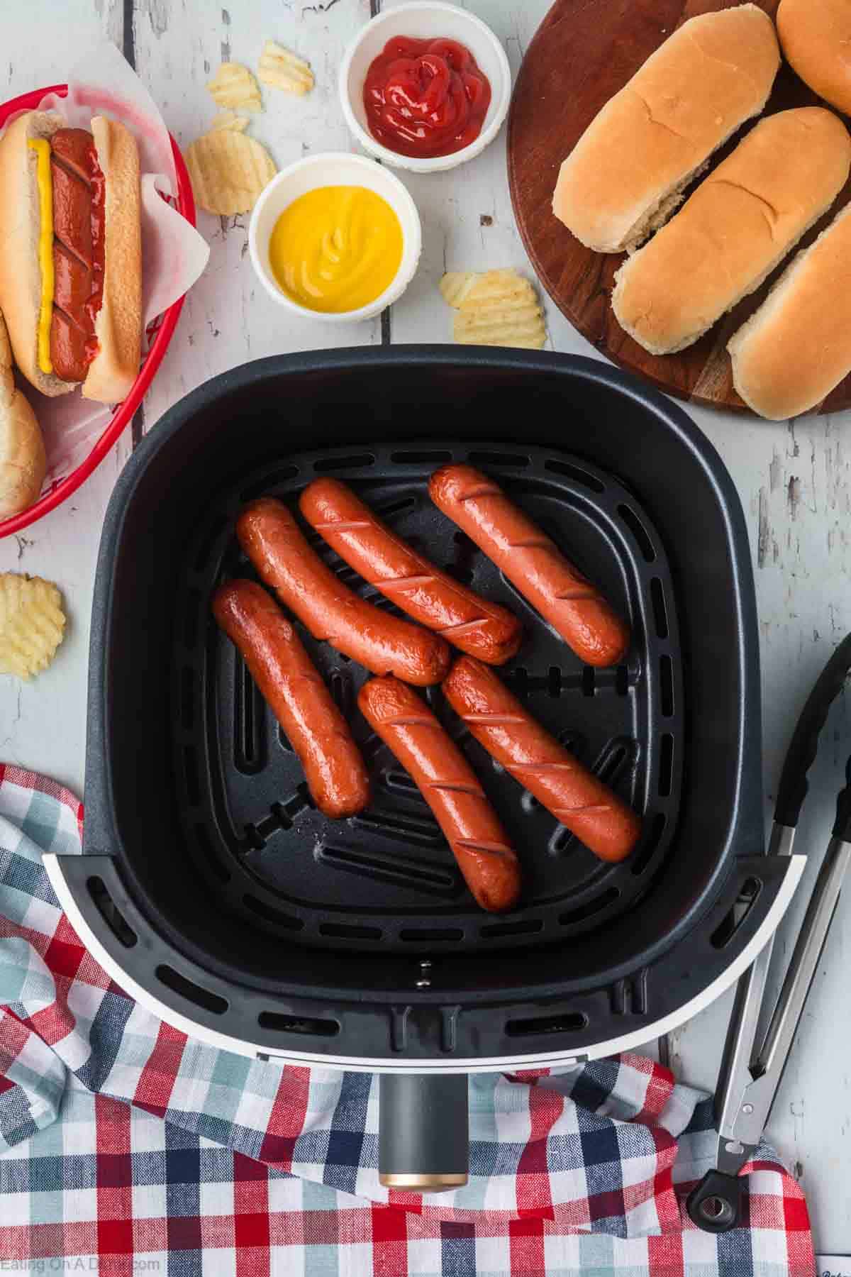 Cooked hot dogs in an air fryer basket with hot dogs in a basket with bowls of mustard and ketchup on the side with a plate of hot dog buns