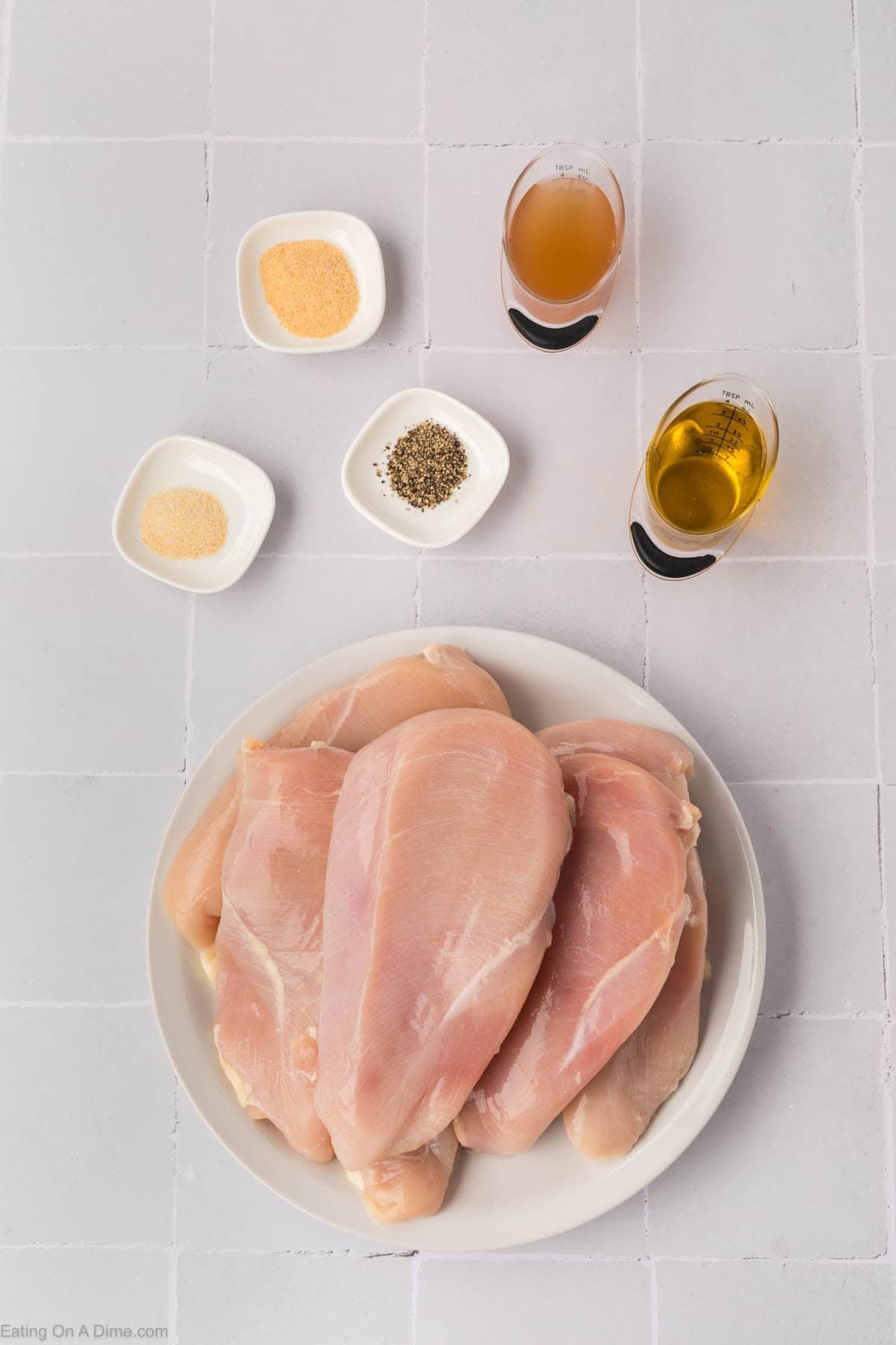 Plate of raw chicken breasts on a white tiled surface, ready for a lesson on how to grill chicken breasts, surrounded by small bowls of seasoning like garlic powder, onion powder, and pepper. Two glasses contain liquids, possibly oil and vinegar.