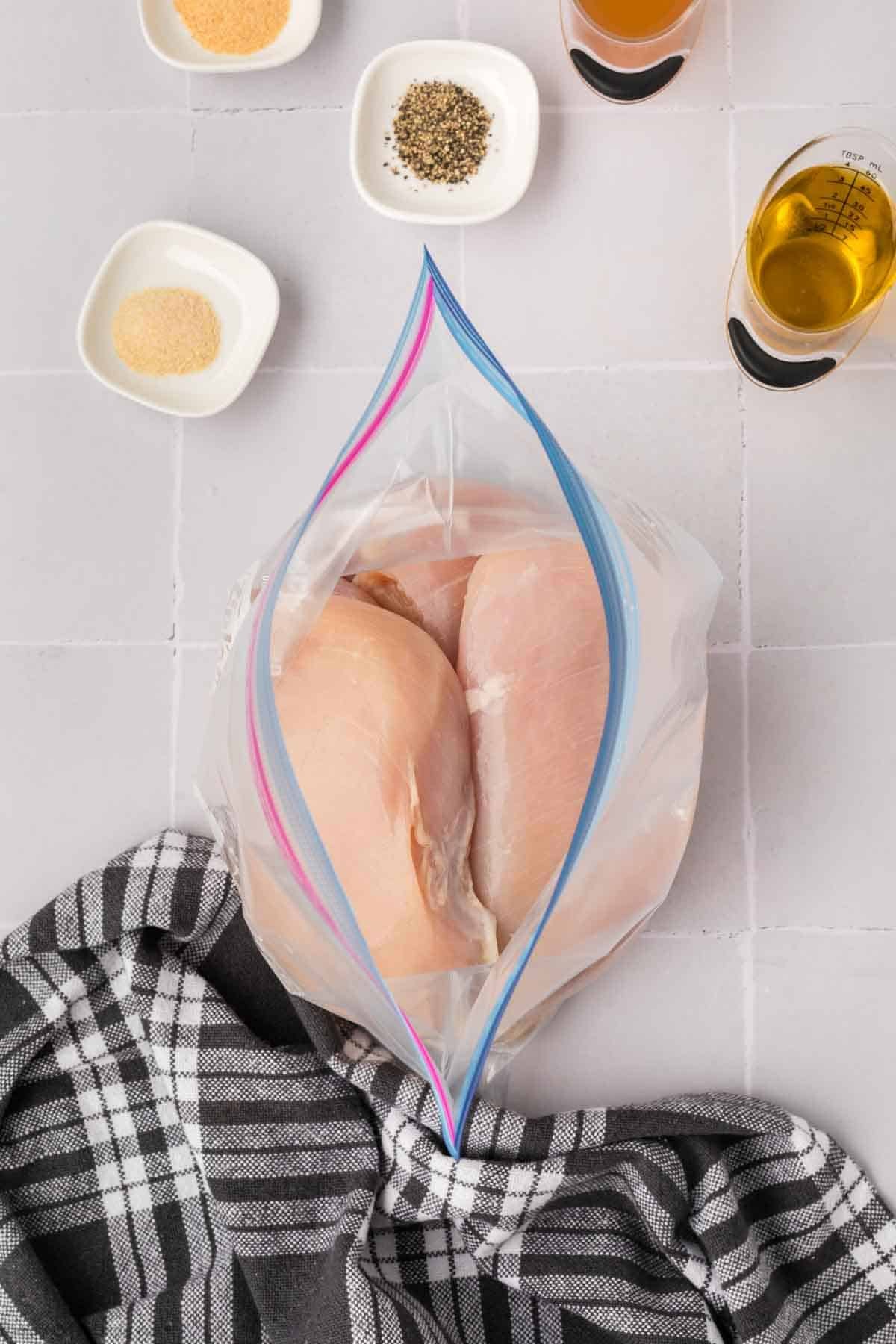 Two raw chicken breasts in a sealed plastic bag, ready for grilling, are surrounded by spices and seasonings on small plates, a measuring cup with oil, and a checkered kitchen towel on the tiled surface.