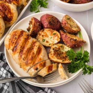 A plate featuring expertly grilled chicken breast and seasoned roasted red potatoes, garnished with parsley, showcases the essence of how to grill chicken breasts to perfection. A fork slices through the succulent meat, while a black and white checkered cloth napkin rests nearby.