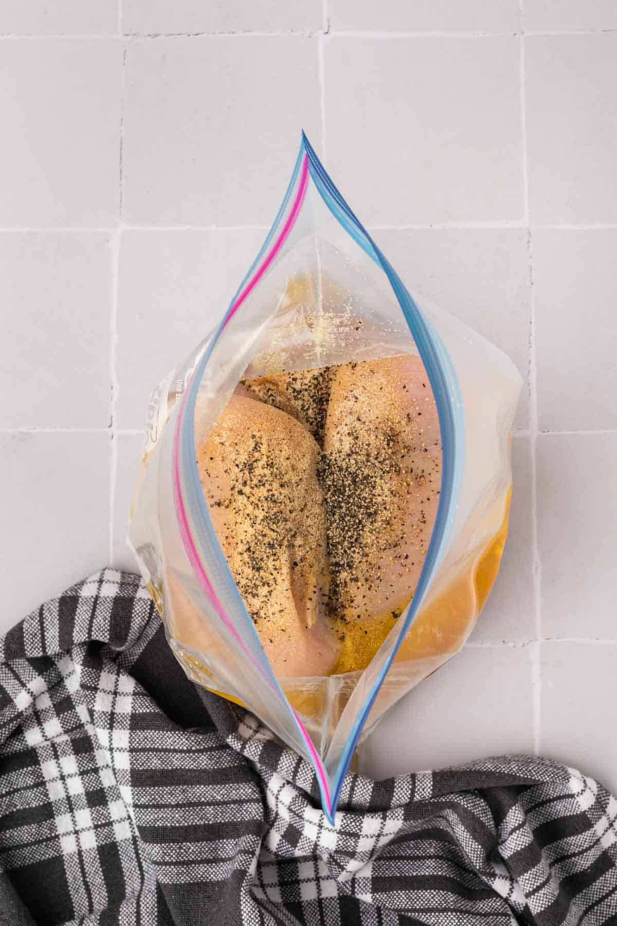 Top-down view of a plastic resealable bag with two chicken breasts seasoned with black pepper, ideal for those wondering how to grill chicken breasts perfectly. They're placed on a light tile surface, with a black and white checkered cloth lying beside the bag.