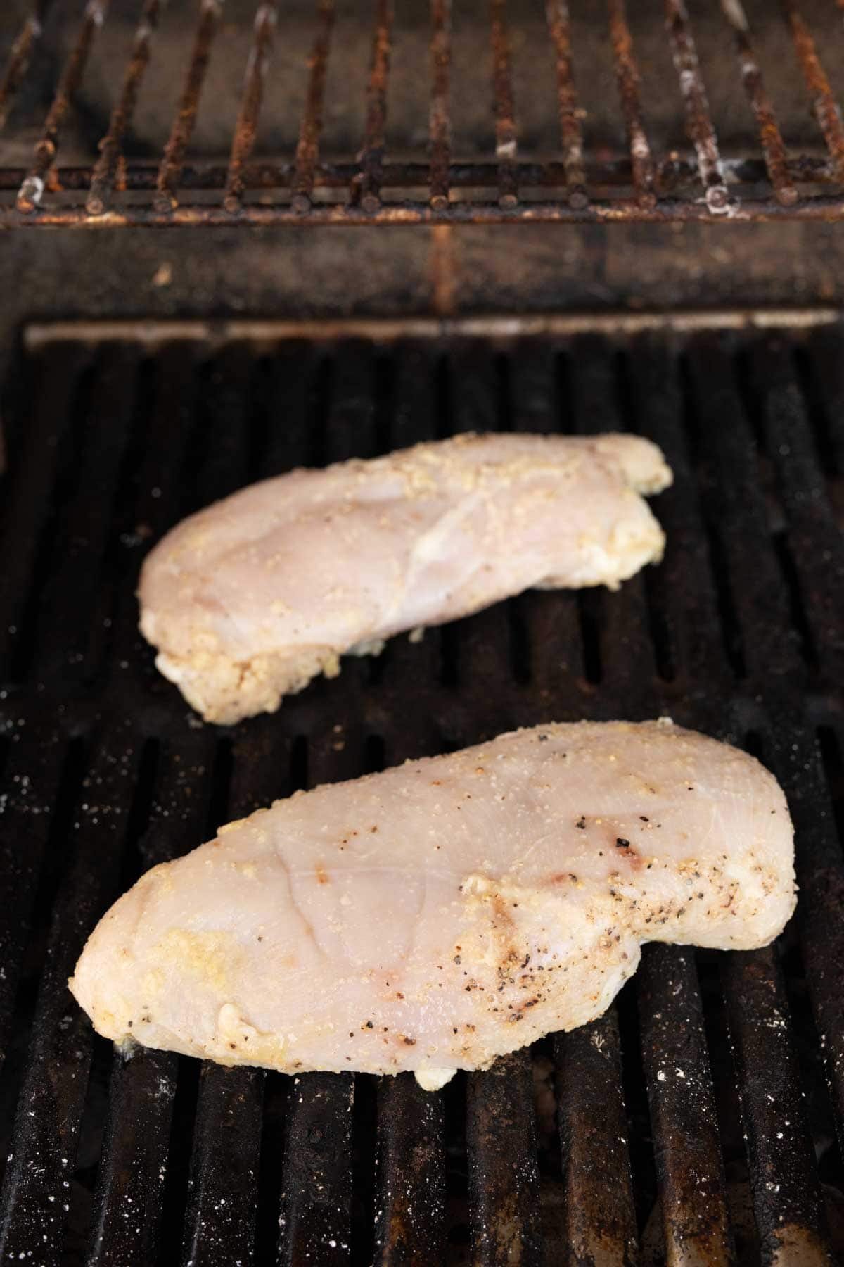 Two uncooked chicken breasts are placed on a grill with visible black grates, showing the perfect guide on how to grill chicken breasts. The chicken is lightly marinated and seasoned, ready for grilling. In the background, the upper grate appears unused.