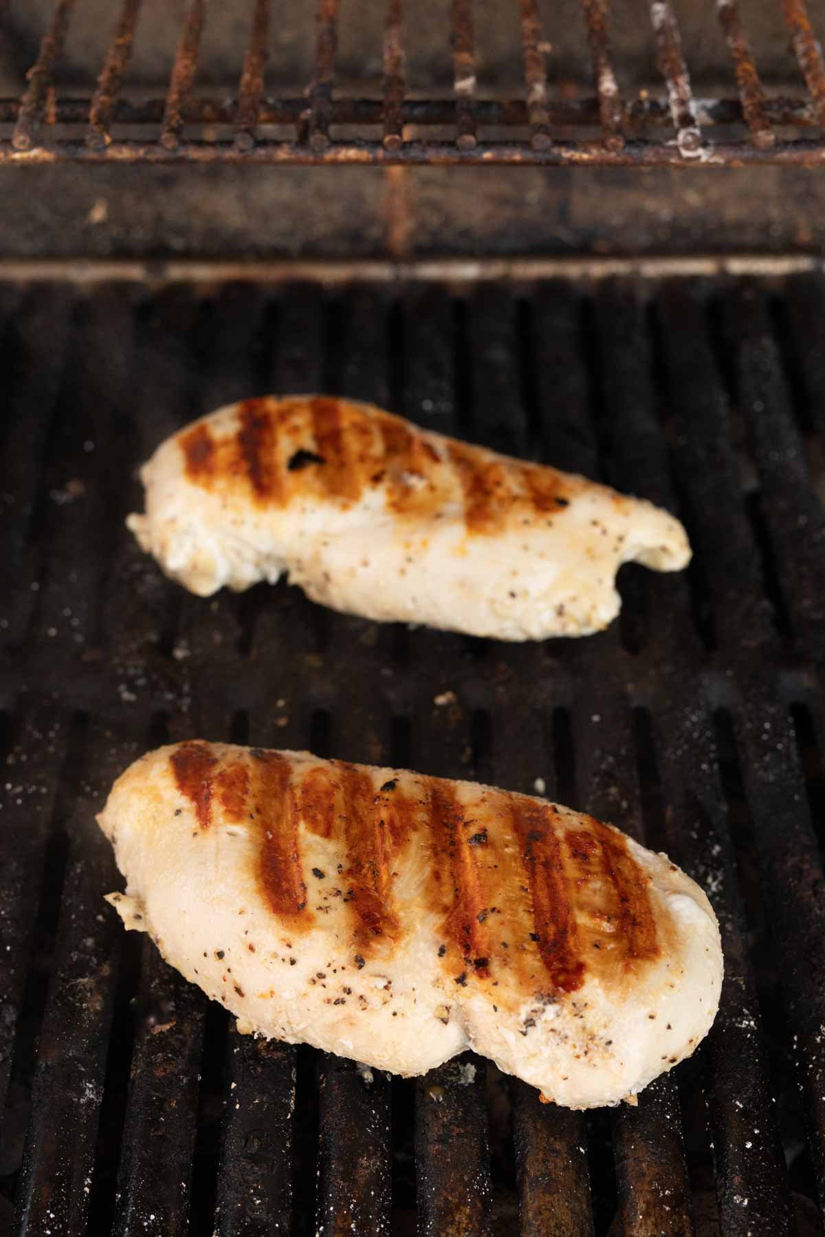 Two seasoned chicken breasts sizzle on a black barbecue grate, showcasing how to grill chicken breasts perfectly. With visible char marks and a blend of white and golden brown hues, the slightly weathered grill adds charm to the partially cooked masterpiece.