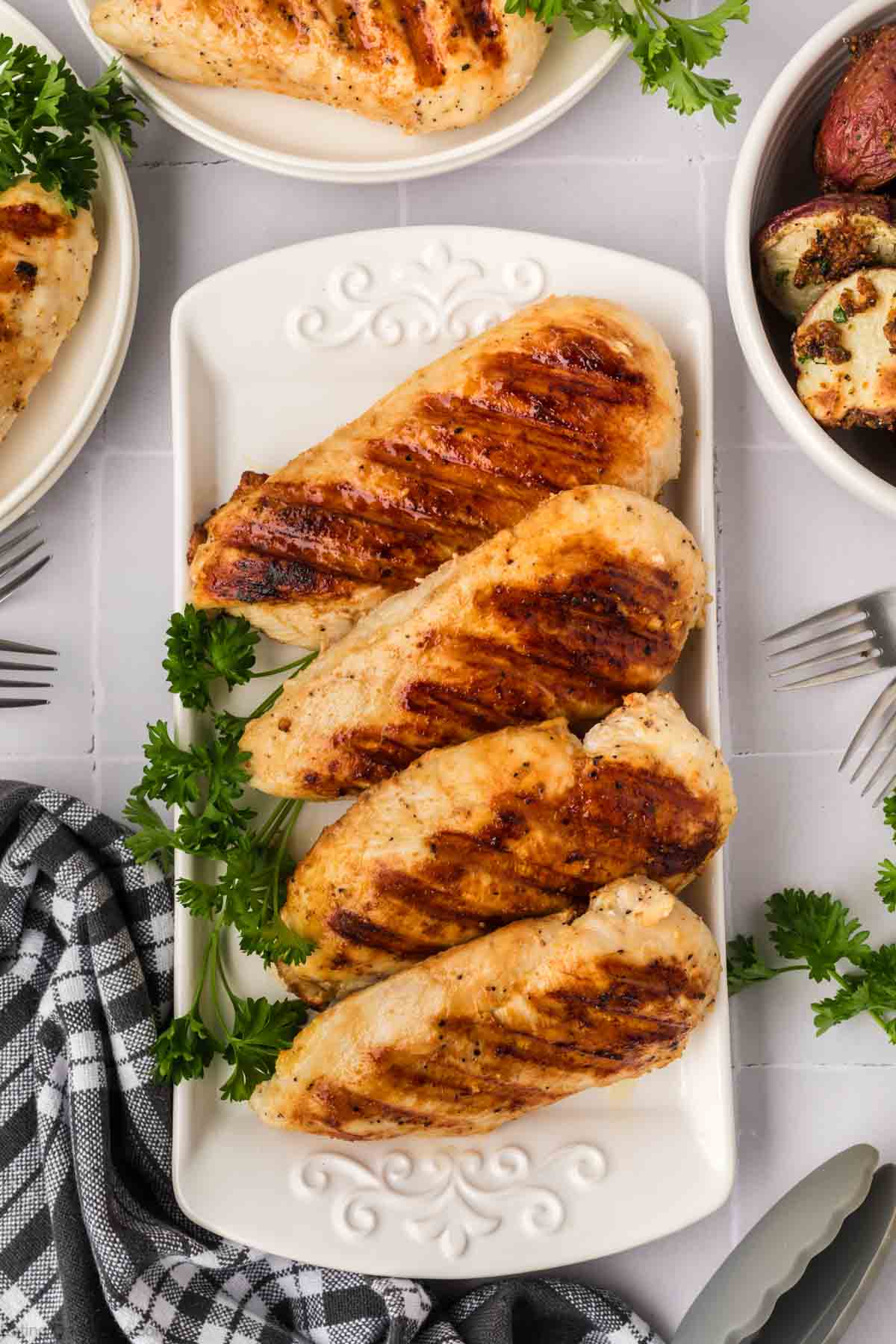 Delicious grilled chicken breasts are elegantly arranged on a white platter, garnished with parsley. Surrounded by checkered napkins, forks, and additional dishes like roasted potatoes, this inviting setup offers tips on how to grill chicken breasts perfectly every time. The scene is set on a white, tiled surface.