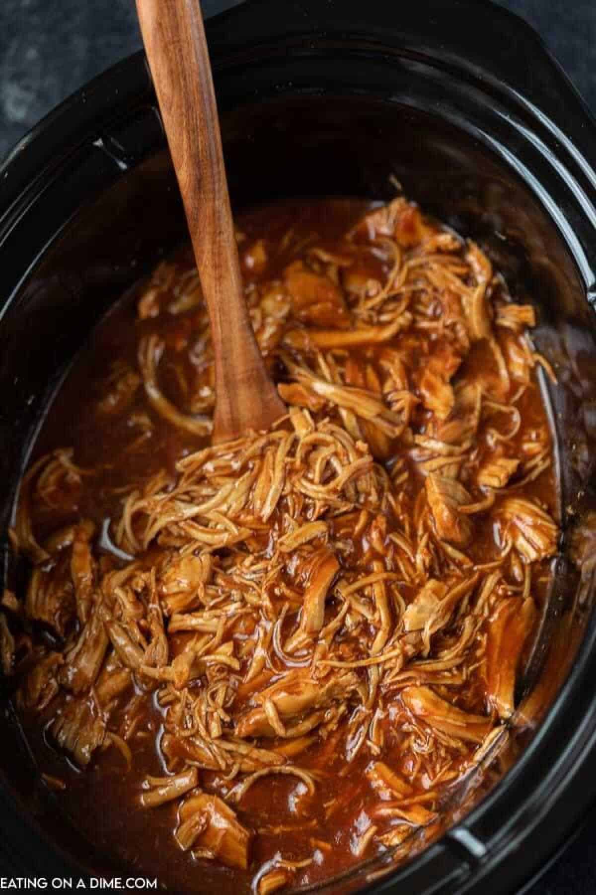 BBQ pulled chicken in a black crock pot with a wooden spoon in the chicken mixture. 