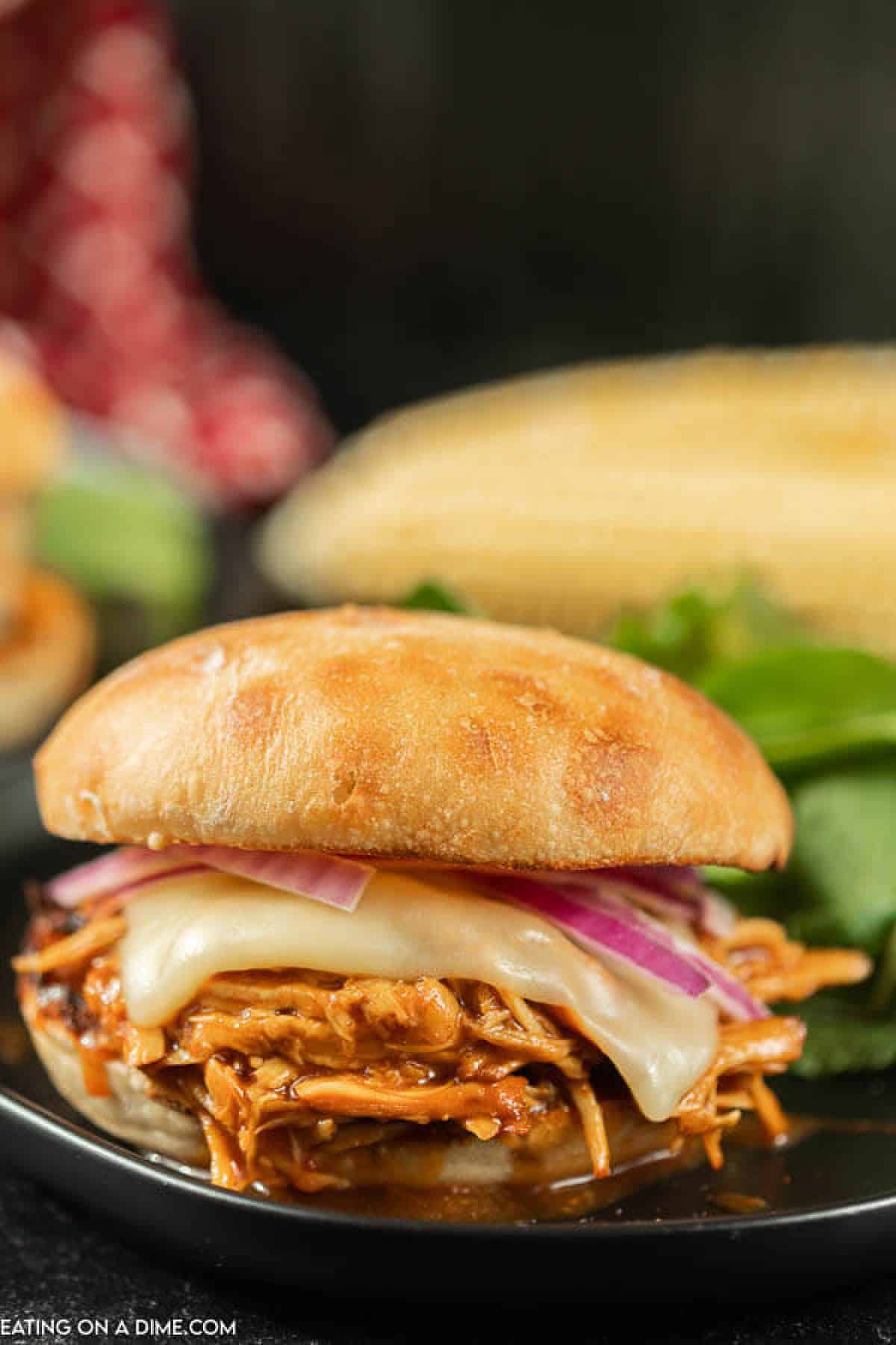 A bbq pulled chicken sandwich topped with red onion and cheese with corn in the back ground 