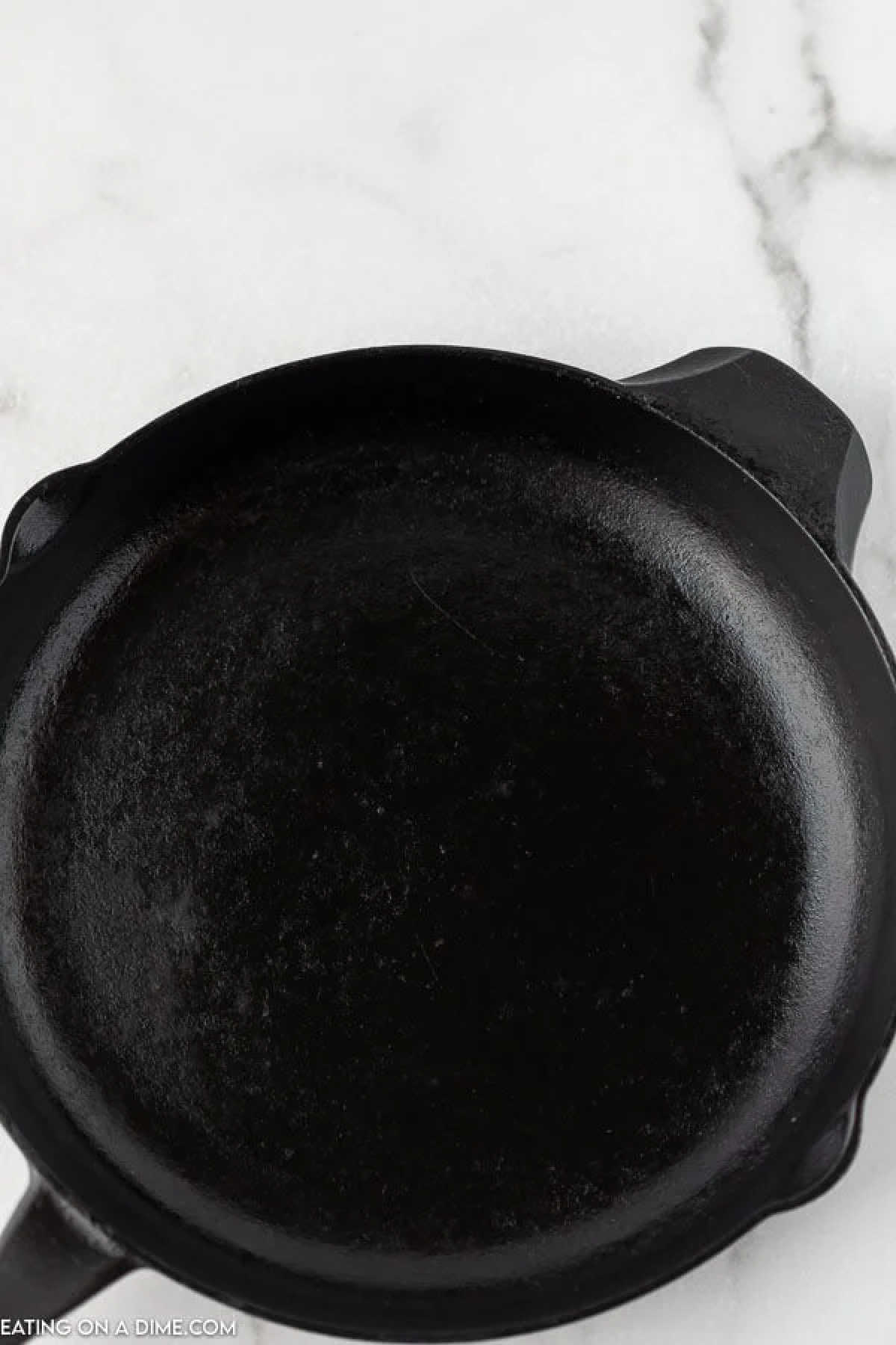 A black cast iron skillet, expertly seasoned, rests on a white marble surface. With its smooth finish, it's a testament to knowing how to season a cast iron skillet properly.