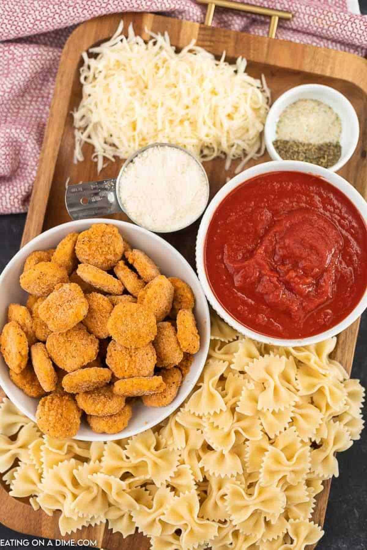 A wooden tray holds a spread of ingredients: a bowl of breaded chicken nuggets, a measuring cup of grated Parmesan cheese, shredded mozzarella, a bowl of marinara sauce, a bowl of uncooked bow tie pasta, and a small dish of Italian seasoning and salt—all you need for the perfect chicken nugget casserole.