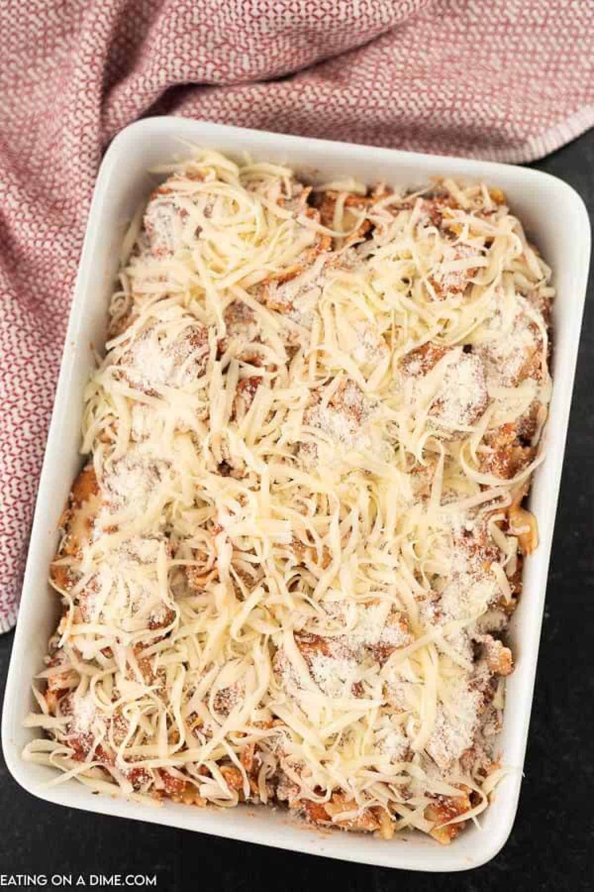 A rectangular baking dish filled with a layered chicken nugget casserole topped with shredded cheese, ready for baking. A red-and-white patterned cloth is partially seen in the background, hinting at a cozy kitchen setting. The dish appears to promise a comforting and delicious meal.