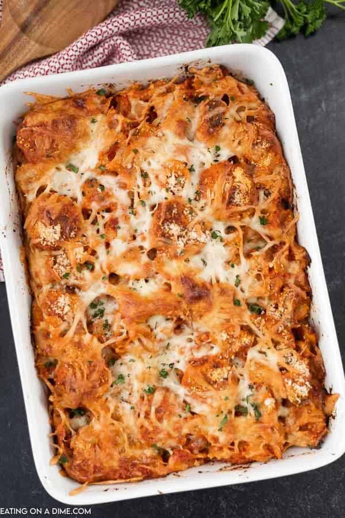 A white baking dish containing a freshly baked chicken nugget casserole topped with golden-brown melted cheese and sprinkled with herbs. The edges are slightly crispy, and the casserole looks hearty and delicious. The dish is placed on a dark surface with a red cloth nearby.