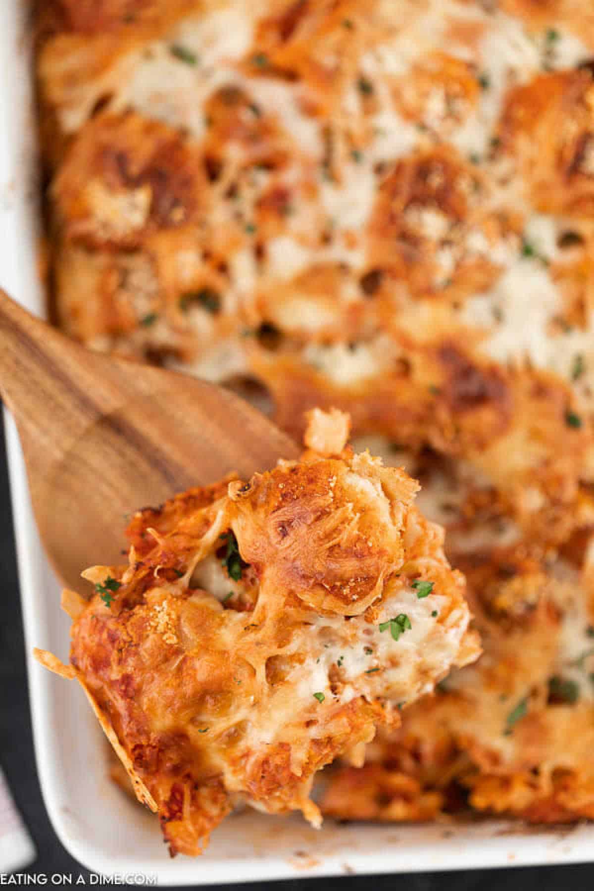 A close-up of a serving of baked chicken nugget casserole with melted cheese being scooped out of a white casserole dish with a wooden spoon. The top is golden brown and garnished with herbs.