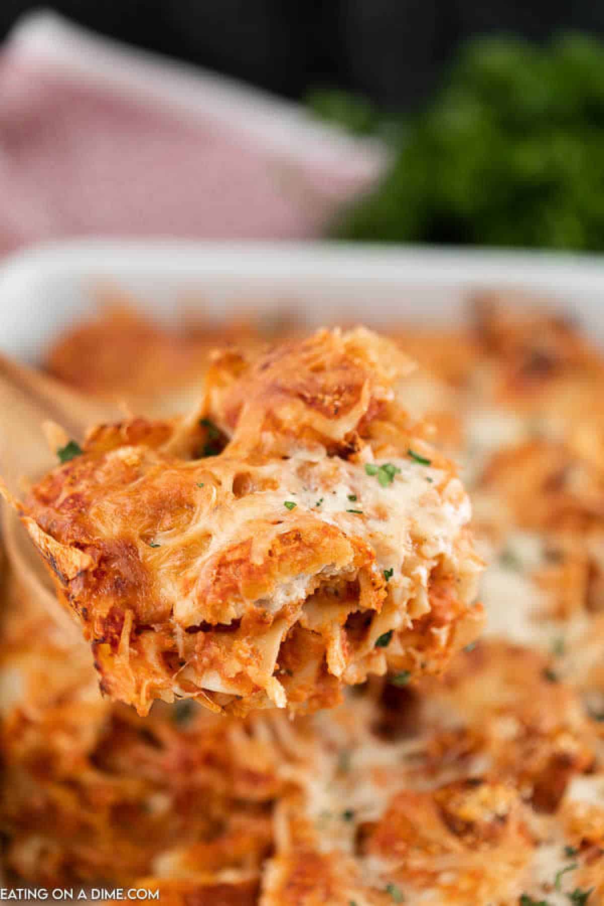 A close-up of a wooden spoon lifting a portion of cheesy baked ziti from a chicken nugget casserole dish. The pasta is covered in melted cheese and garnished with chopped herbs. A blurred background includes a hint of green, possibly fresh herbs or a salad.