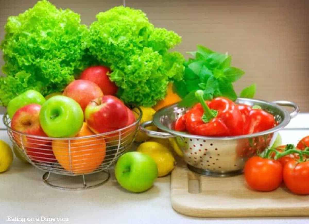 A kitchen counter with a wire basket of apples, oranges, and leafy greens. Next to it, a colander—fresh from a fruit and vegetable wash—holds red bell peppers and basil on a cutting board with tomatoes. Lemons are scattered nearby.