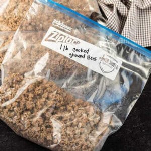 A clear Ziploc bag labeled "1 lb cooked ground beef" contains perfectly cooked meat, ready for storage. Sealed properly to ensure freshness on a dark surface, this is an ideal example of how to freeze ground beef efficiently. A checkered cloth adds a cozy touch in the background.