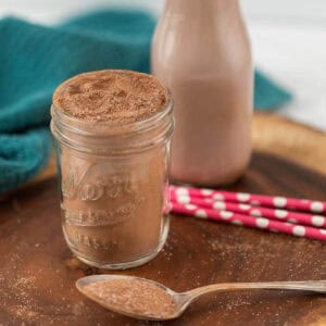 A jar filled with cocoa powder, resembling homemade Nesquik, sits on a wooden surface next to a bottle of chocolate milk. Pink polka-dotted straws and a blue cloth add charm in the background, while a spoon laden with cocoa powder lies in the foreground.