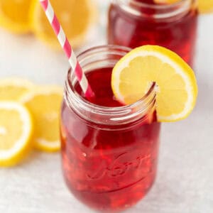 A mason jar brimming with red iced tea, reminiscent of a Starbucks passion tea lemonade recipe, is garnished with a lemon slice on the rim and features a pink and white striped straw. In the background, another jar and several lemon slices are scattered on a light surface.