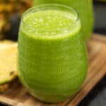 A close-up of a glass filled with a vibrant green spinach smoothie on a wooden surface. The frothy texture is inviting, and a slice of pineapple is partially visible in the background, hinting at the tropical flavors within.