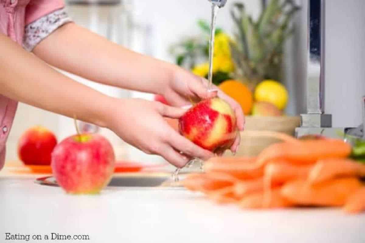 A person uses fruit and vegetable wash while rinsing a red apple under the kitchen faucet. Other apples and freshly washed carrots lie on the counter, set against a backdrop of blurred fruits and flowers.