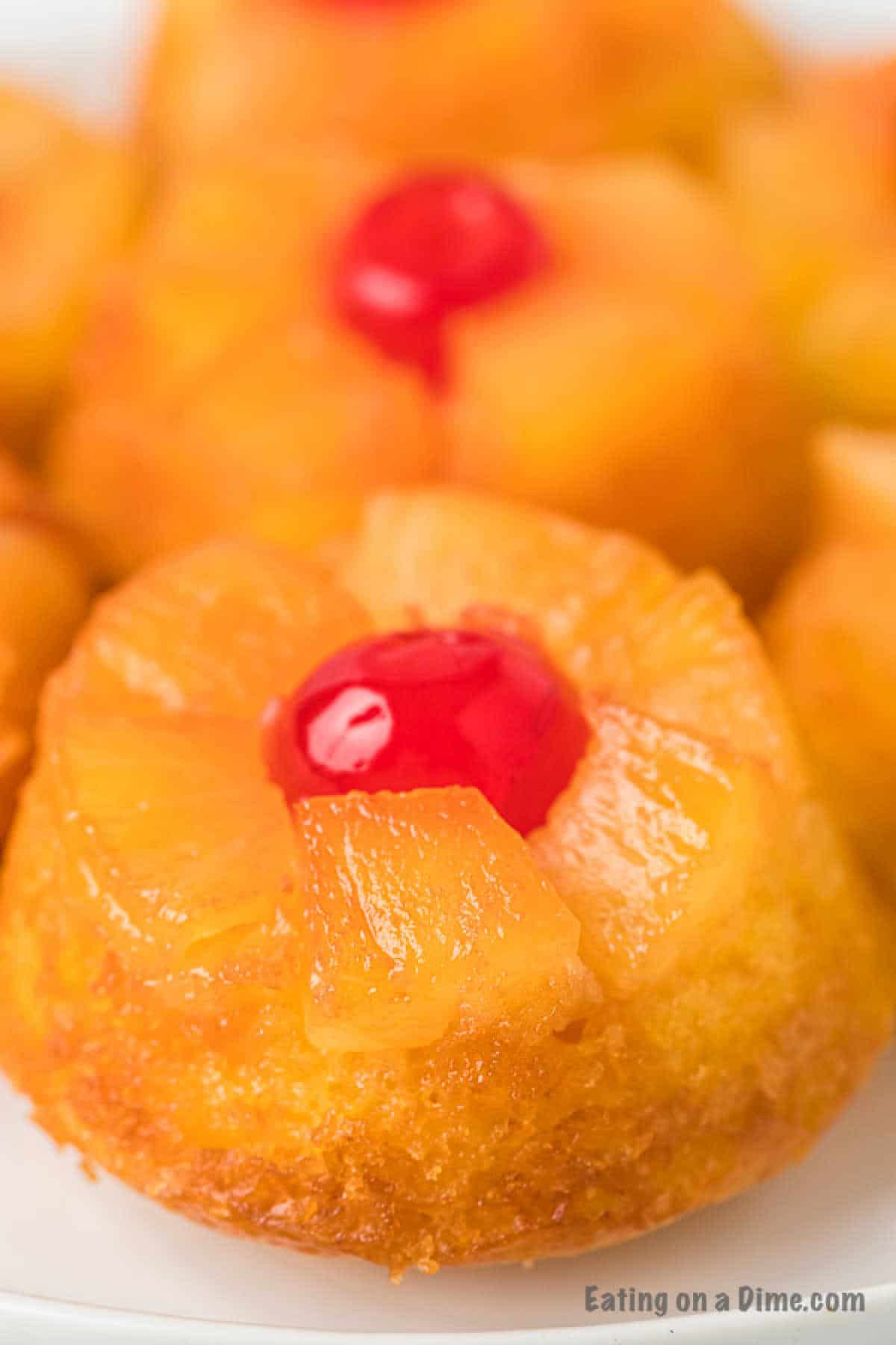 Close-up of mini pineapple upside-down cupcakes adorned with a cherry. The golden-brown cupcakes feature pineapple slices with a vivid red cherry in the center. Their texture looks moist and slightly glossy, while a portion of another cupcake peeks into the background.