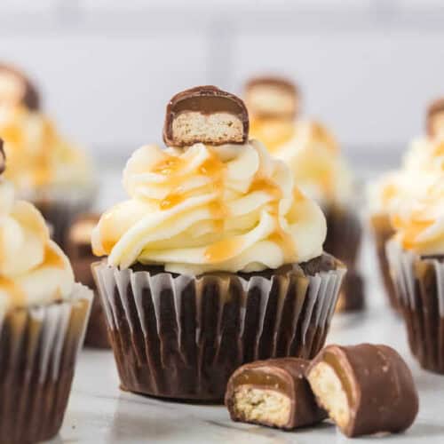 A chocolate cupcake topped with fluffy cream frosting, drizzled with caramel sauce, is crowned by a piece of Twix bar filled with biscuit. Another split in half rests beside the cupcake, while more Twix Bars cupcakes are artfully blurred in the background.