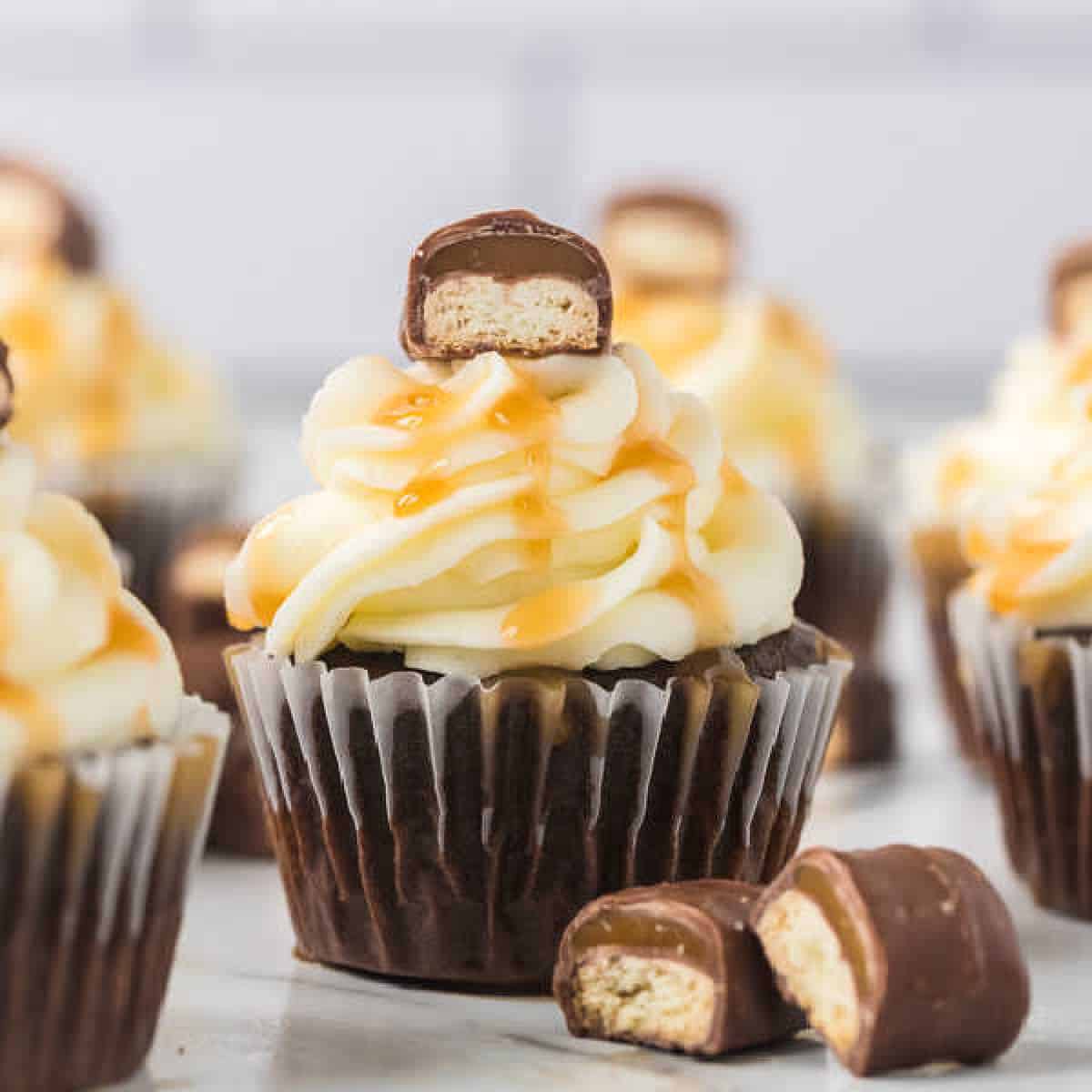 A chocolate cupcake topped with fluffy cream frosting, drizzled with caramel sauce, is crowned by a piece of Twix bar filled with biscuit. Another split in half rests beside the cupcake, while more Twix Bars cupcakes are artfully blurred in the background.