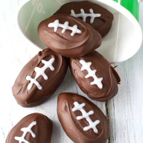 Football-shaped chocolate peanut butter balls spill out of a green and white container, each treat decorated with white icing to mimic football laces. They rest elegantly on a light wooden surface, capturing the spirit of the game in every delicious bite.