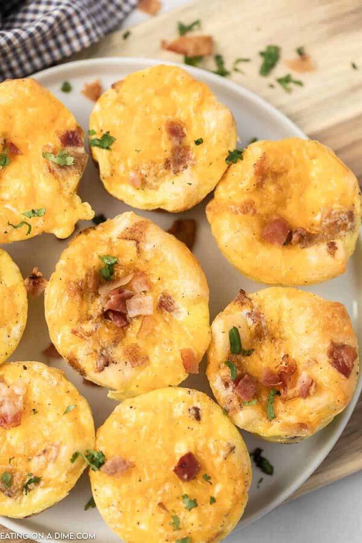 A plate of eight golden breakfast egg muffins topped with bits of bacon and sprinkled with herbs. They are neatly arranged on a white plate, placed on a light wooden surface. A checkered cloth is partially visible in the background.