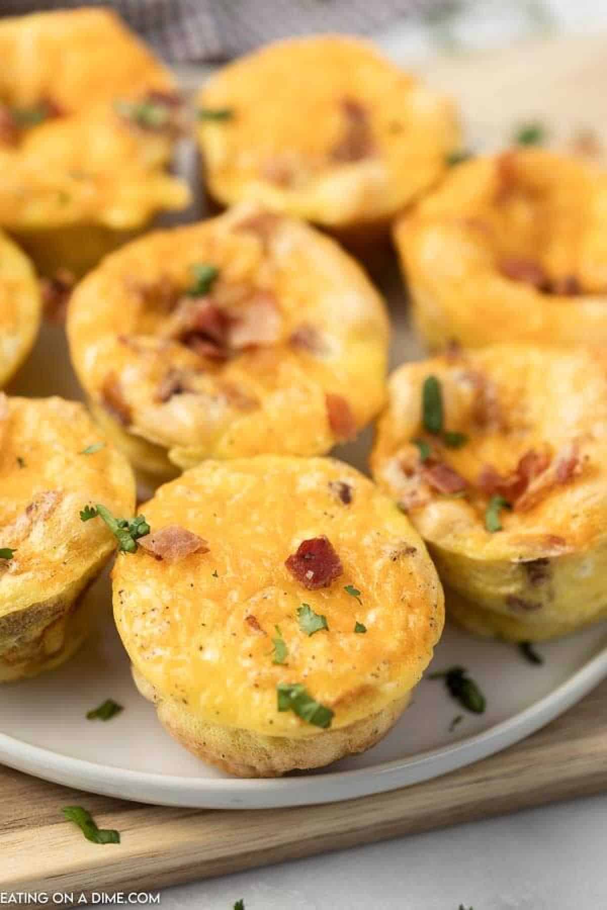 Close-up of a plate with golden brown breakfast egg muffins topped with bacon bits and sprinkled with chopped herbs. The muffins are beautifully arranged on a light-colored platter, inviting you to dive into their deliciousness.