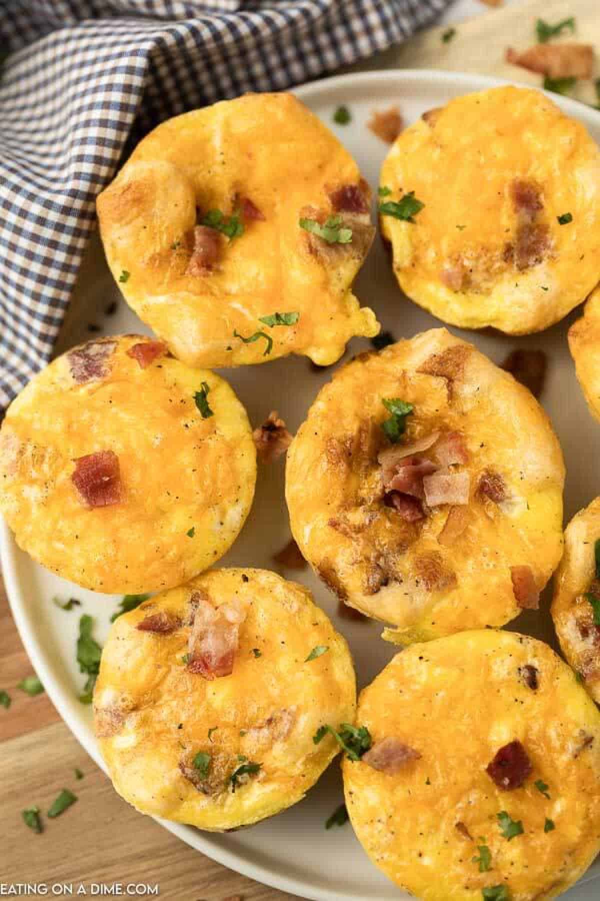 A plate of golden, fluffy breakfast egg muffins garnished with bits of bacon and chopped herbs. A blue and white checkered cloth is partially visible in the background.