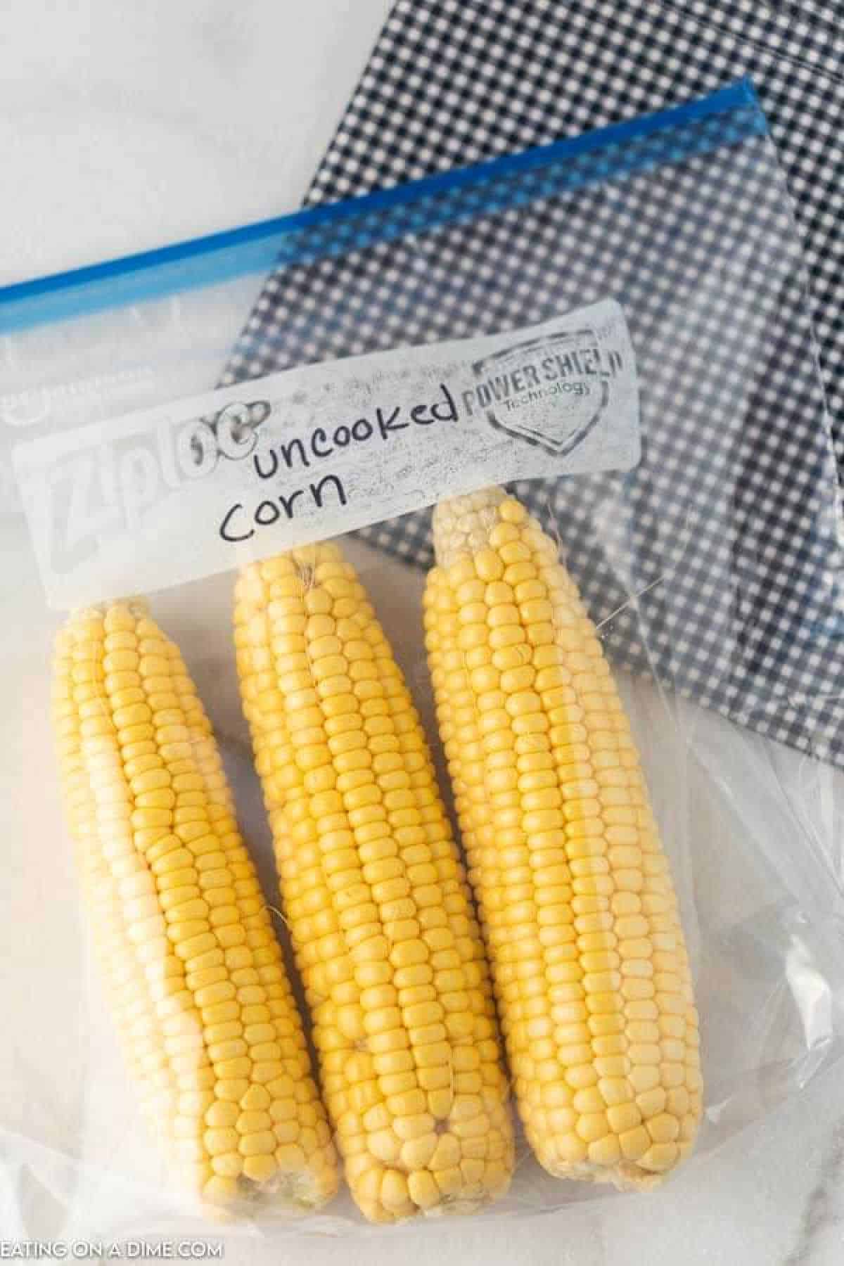 Three uncooked ears of corn rest inside a labeled clear plastic zipper bag, ready for storage. A black and white checkered cloth is partially visible in the background, adding a touch of charm. Perfect for anyone learning how to freeze corn on the cob efficiently and stylishly.