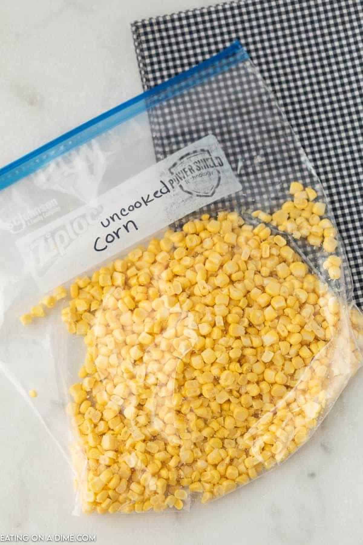 A clear ziplock bag labeled "uncooked corn" is filled with yellow kernels, ready for those learning how to freeze corn on the cob. The bag sits on a black and white checkered surface, suggesting simplicity and ease in preservation.