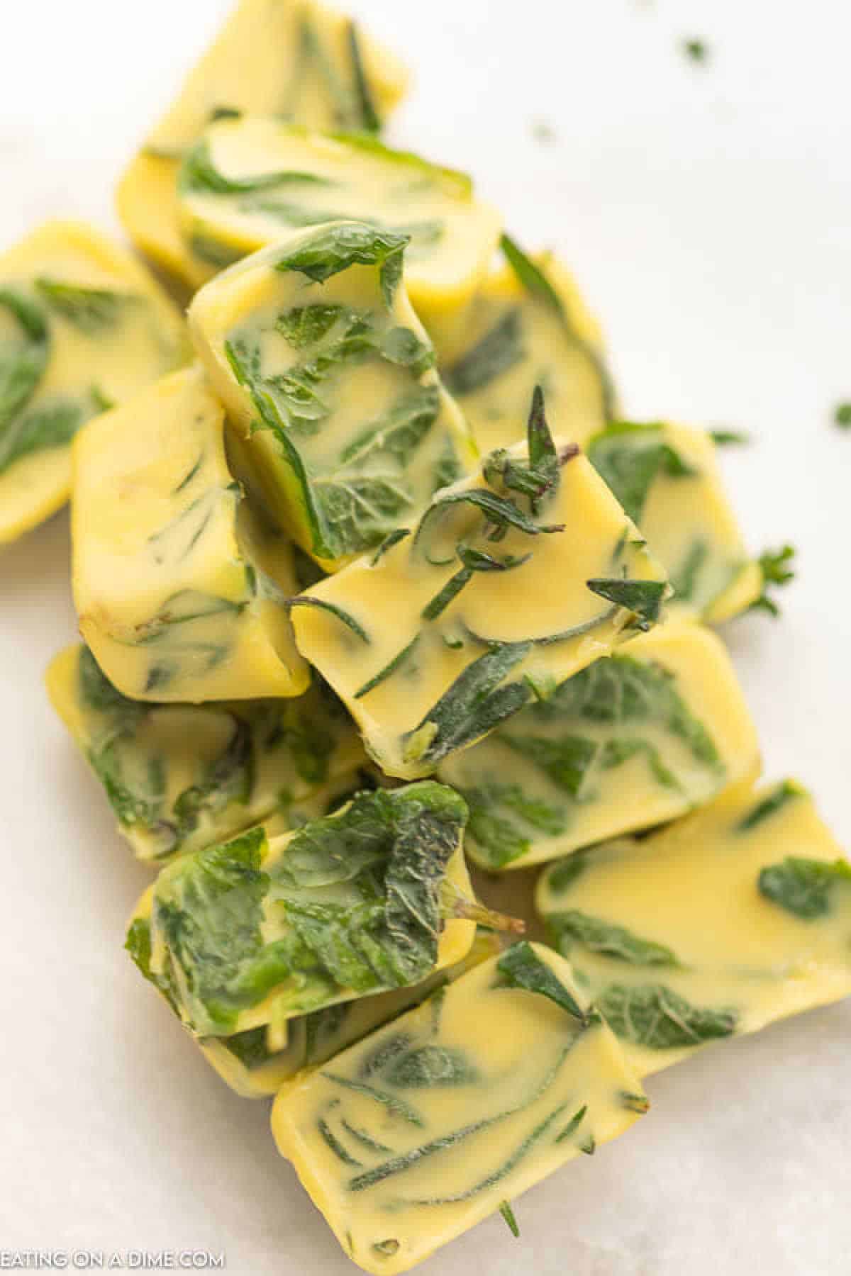 Cubes of herb-infused butter, with visible green leaves, are stacked on a white background. The butter appears creamy and smooth, evenly distributing herbs throughout each cube. Discover how to freeze herbs perfectly within these flavorful butter blocks, preserving freshness with ease.