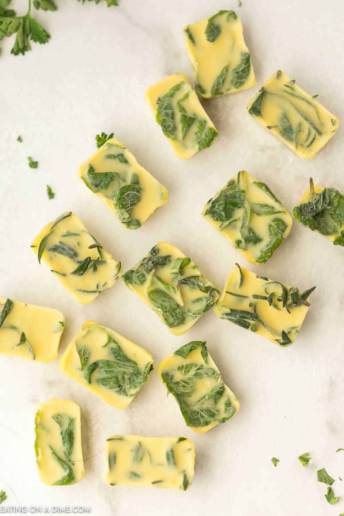 Rectangular blocks of yellow butter mixed with green herbs scattered on a white surface, showcasing an effortless way of how to freeze herbs. Some parsley leaves are visible in the background, adding a fresh touch.