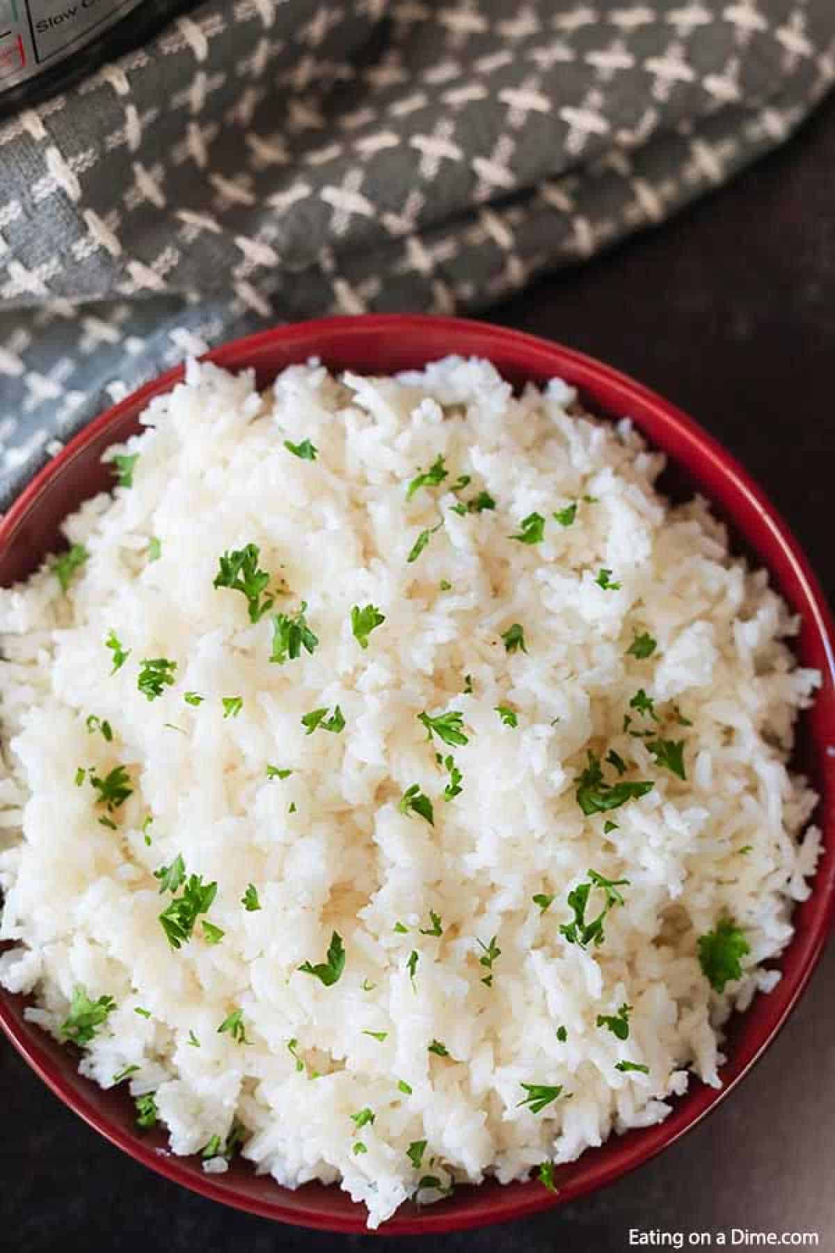 A bowl of fluffy white rice, perfect for exploring ways to reuse leftover rice, is garnished with chopped parsley and sits enticingly on a dark surface. A patterned gray cloth is partially visible in the background, hinting at kitchen creativity.