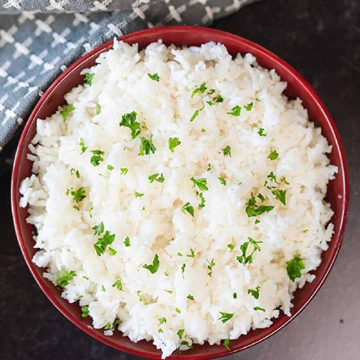 A red bowl filled with fluffy white rice, garnished with finely chopped green herbs, sits on a dark textured surface. A gray and white patterned cloth is partially visible beside the bowl, suggesting creative ways to reuse leftover rice for an inviting and delicious meal.