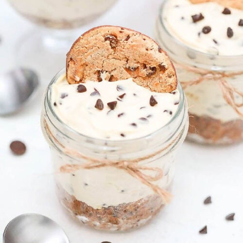 A mini no-bake chocolate chip cookie cheesecake jar dessert with layers of creamy filling and cookie pieces, topped with a cookie and sprinkled with chocolate chips. The jar is tied with a string, and a spoon is nearby on a white background.