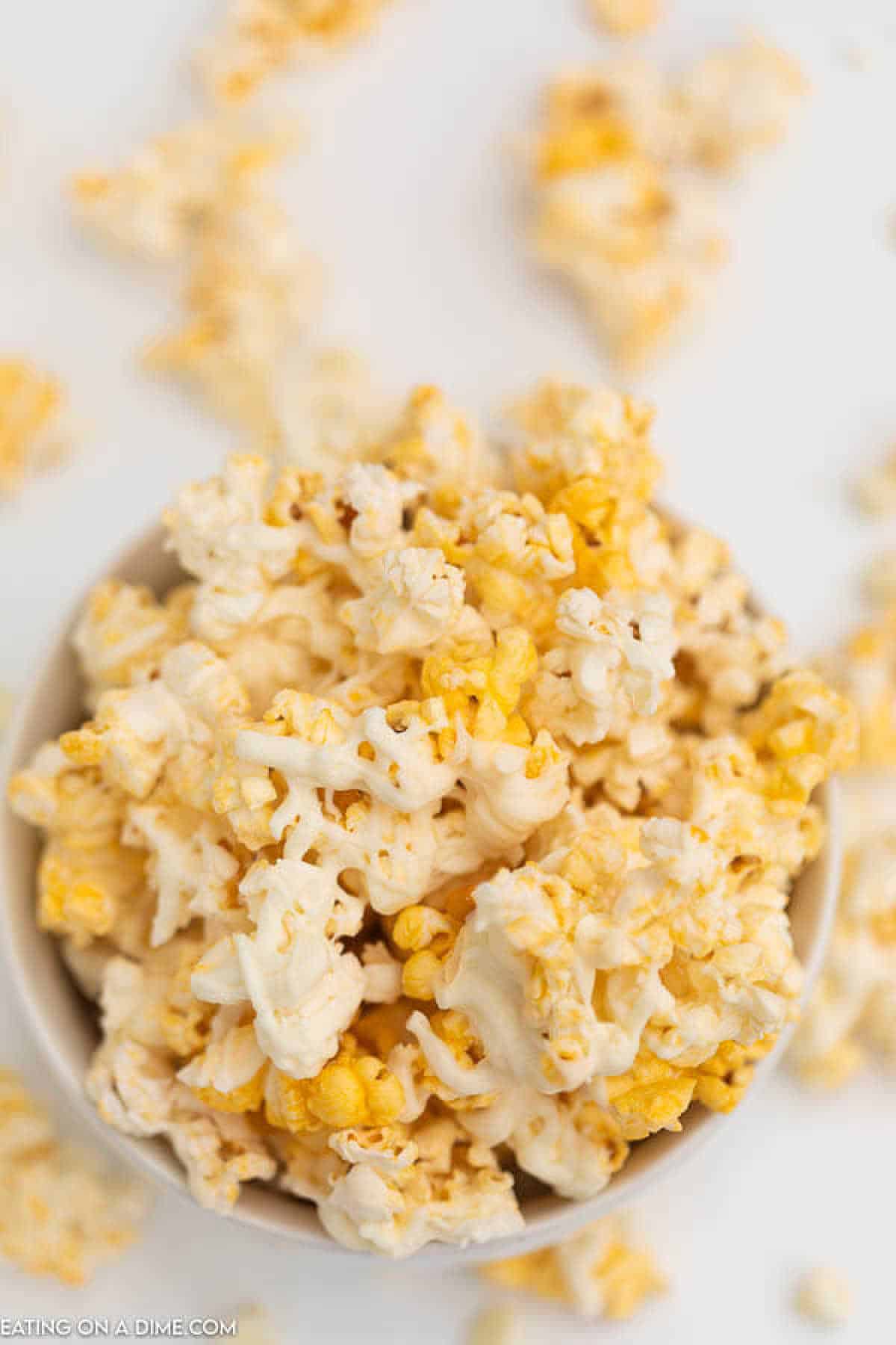 A bowl of popcorn with a generous drizzle of white chocolate rests on a white surface, surrounded by scattered popped kernels. This delightful treat is part of our easy-to-follow white chocolate popcorn recipe, with the golden and white hues creating a striking contrast.