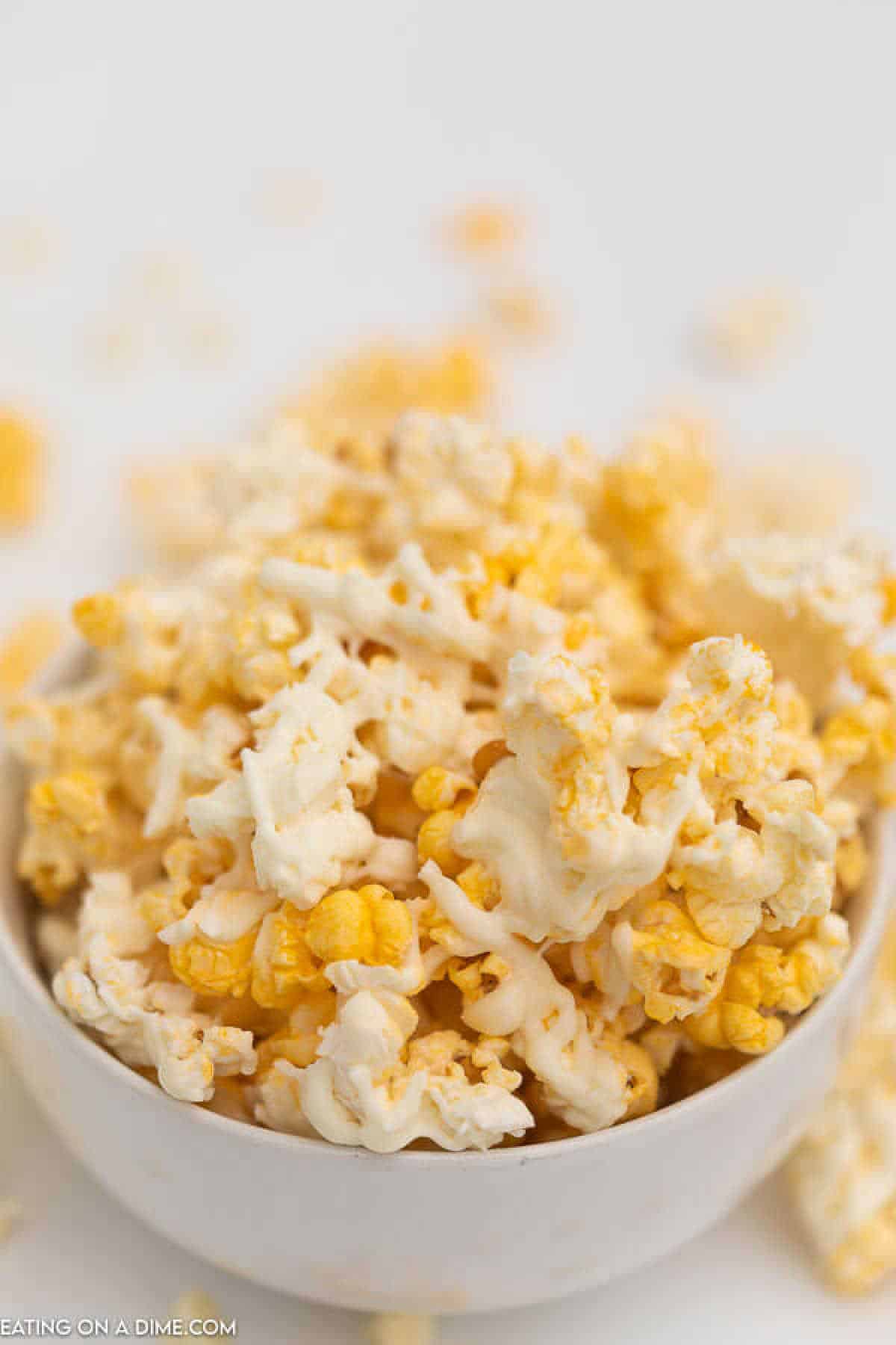 A bowl of popcorn drizzled with a creamy white chocolate coating. The popcorn appears fresh and crunchy, with a few scattered pieces around the bowl. The background is blurred, highlighting the texture and inviting you to explore this delightful white chocolate popcorn recipe.